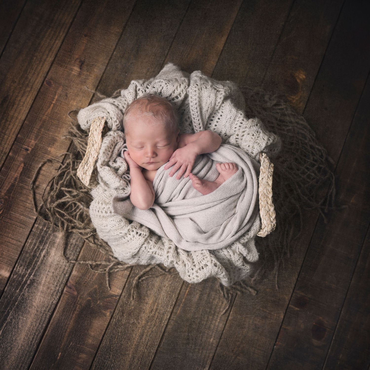 A newborn sleeping peacefully in a cream knit wrap on dark wooden floorboards.