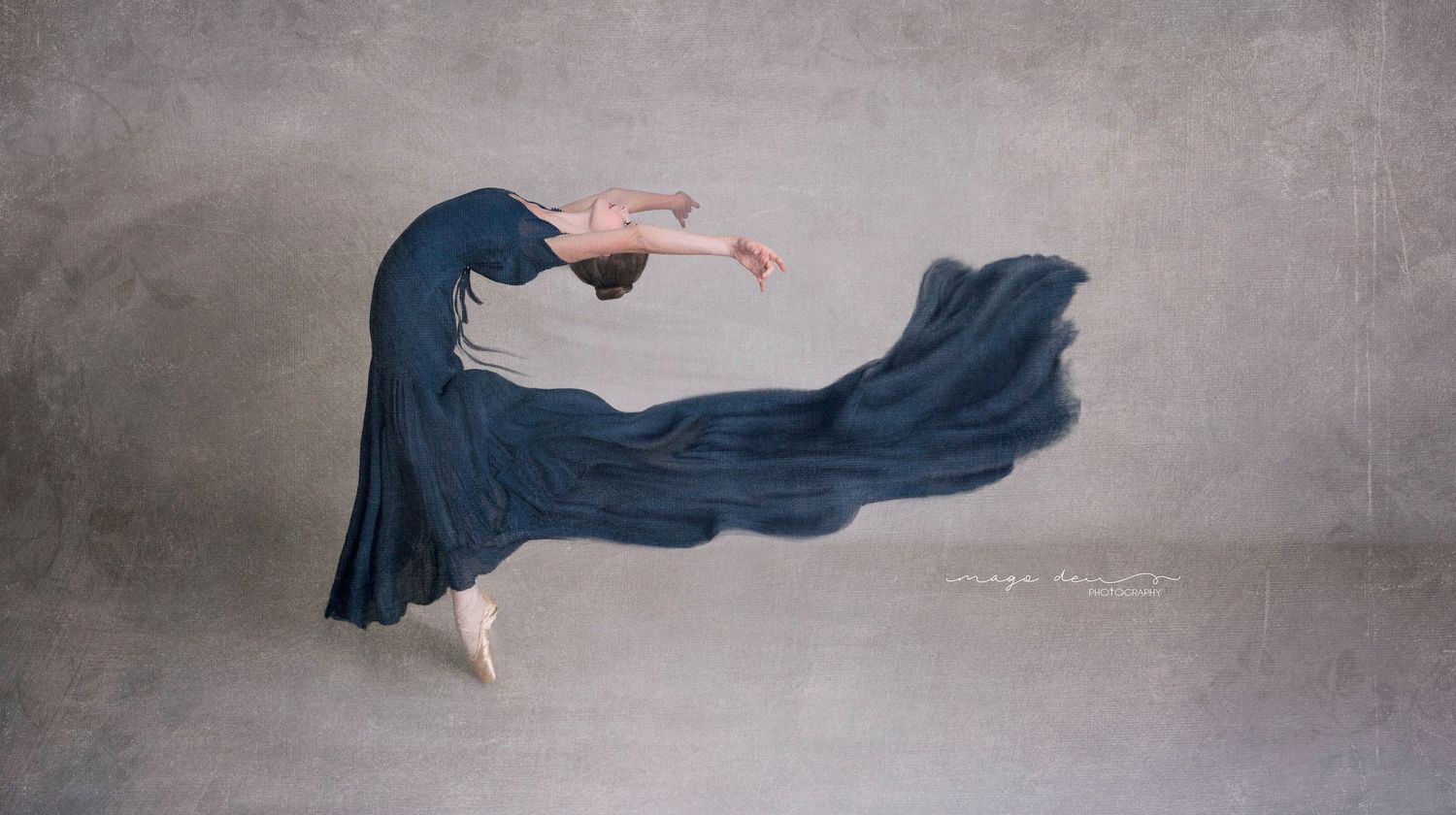 A dancer in a flowing navy blue dress performs a graceful arched movement against a textured gray background.