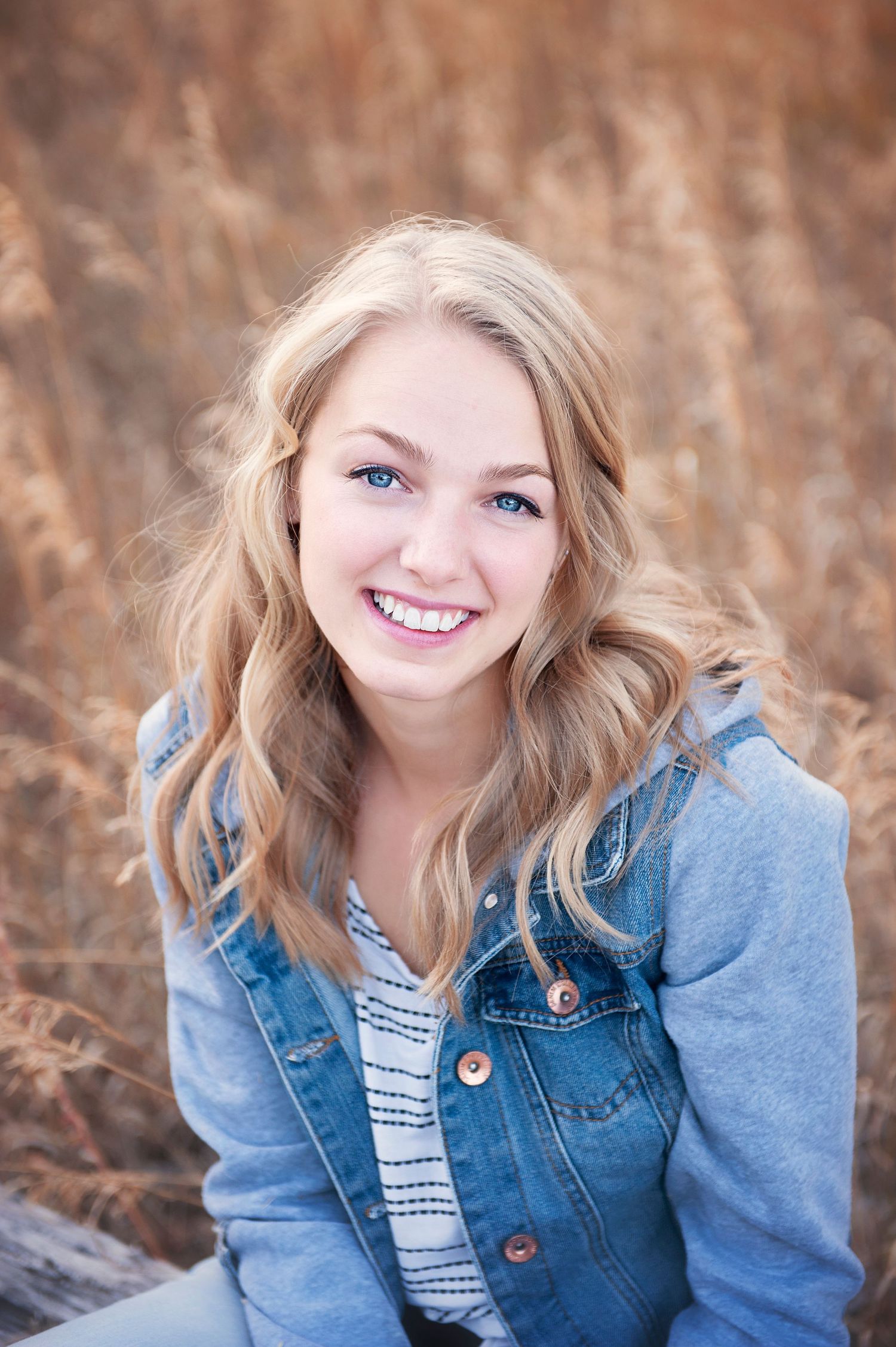 Portrait of a person in a denim jacket and striped top smiling outdoors against a warm autumn background.