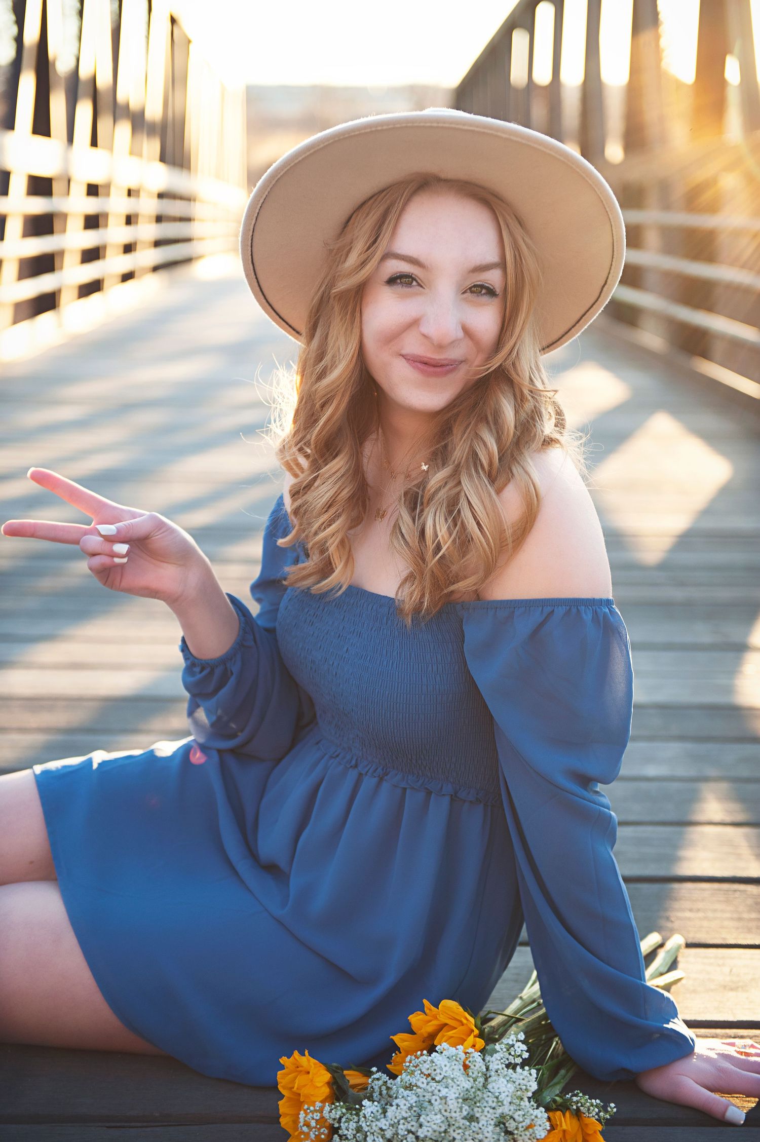 Casual pose on wooden bridge wearing blue off-shoulder dress and wide brim hat.