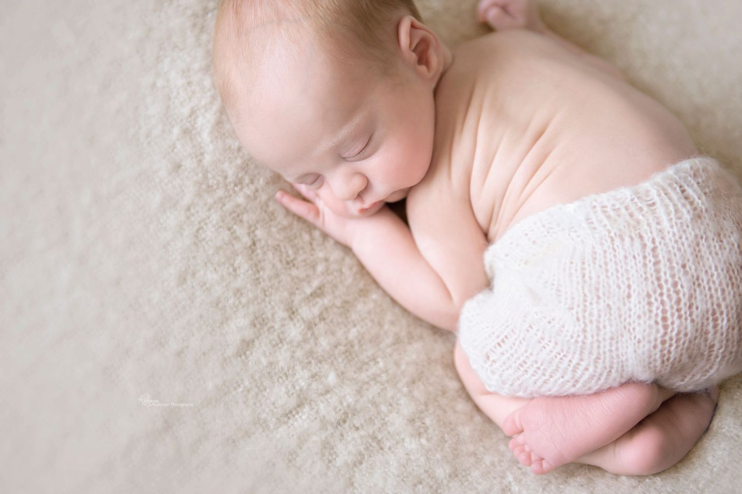 Sleeping newborn wearing white knit wrap lies peacefully on soft beige blanket in studio portrait.