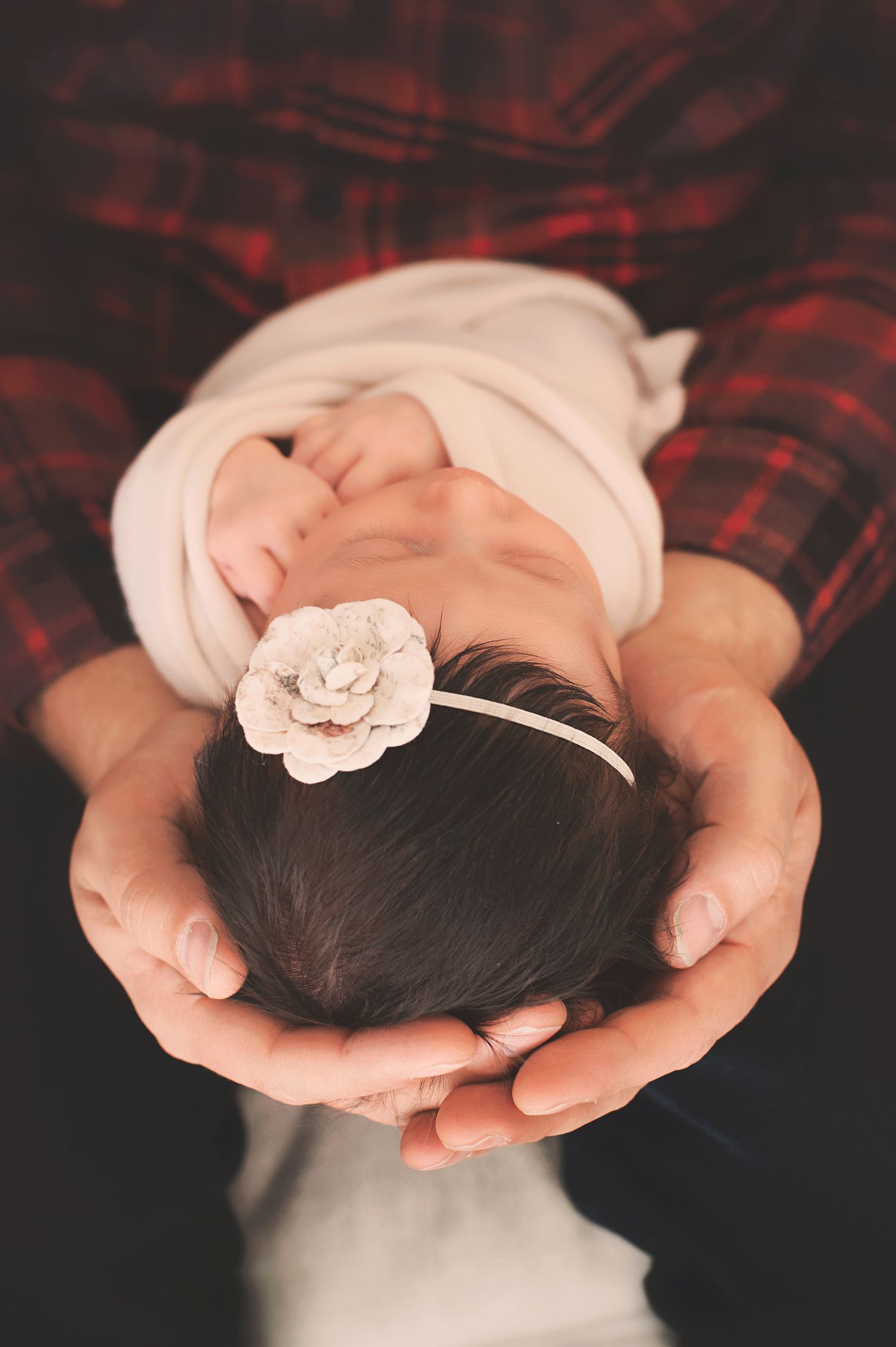 Newborn baby with delicate white flower headband sleeping peacefully in parent's hands.