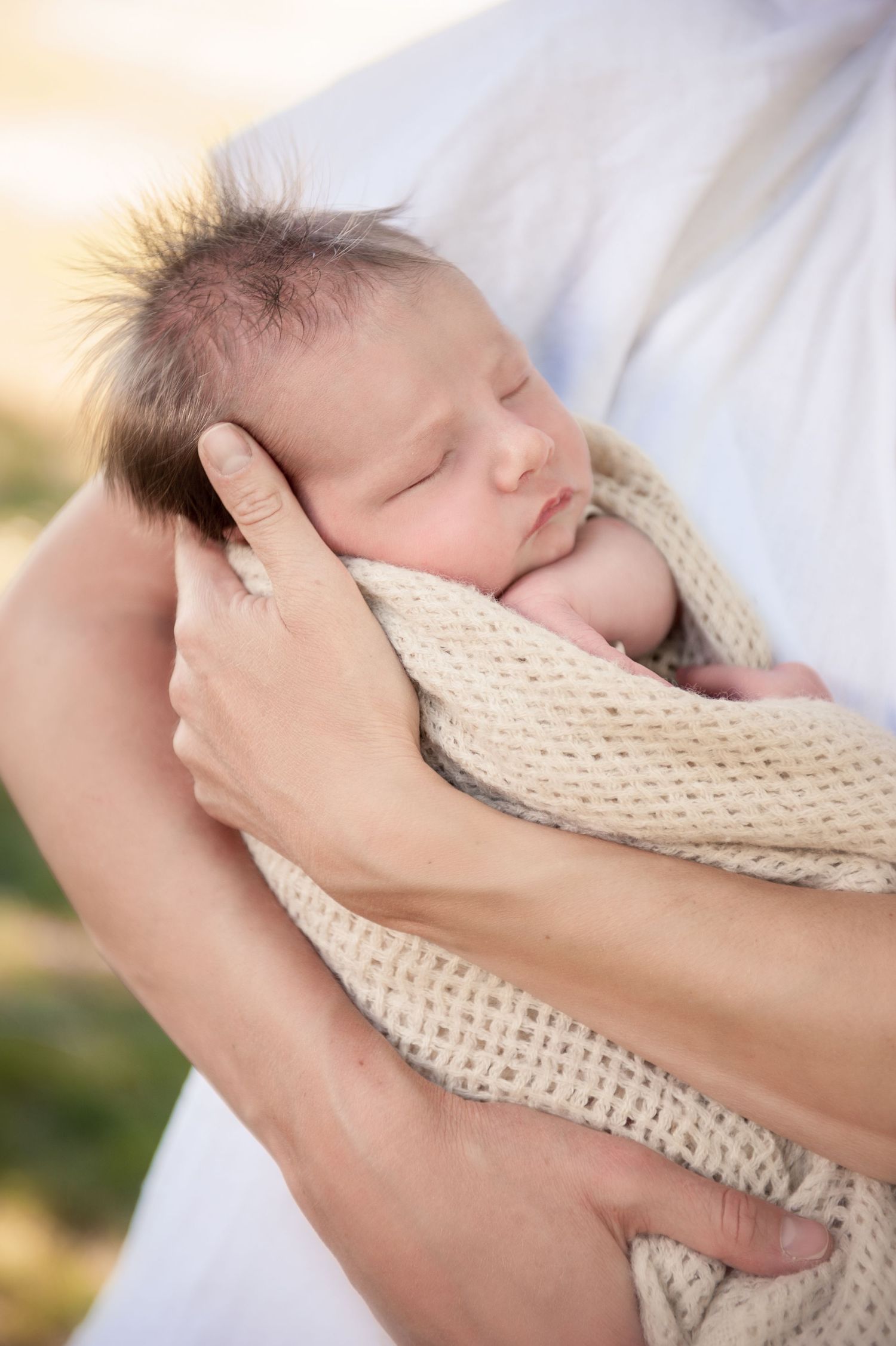 A sleeping newborn wrapped in a cream knit blanket being held gently in sunlit setting.