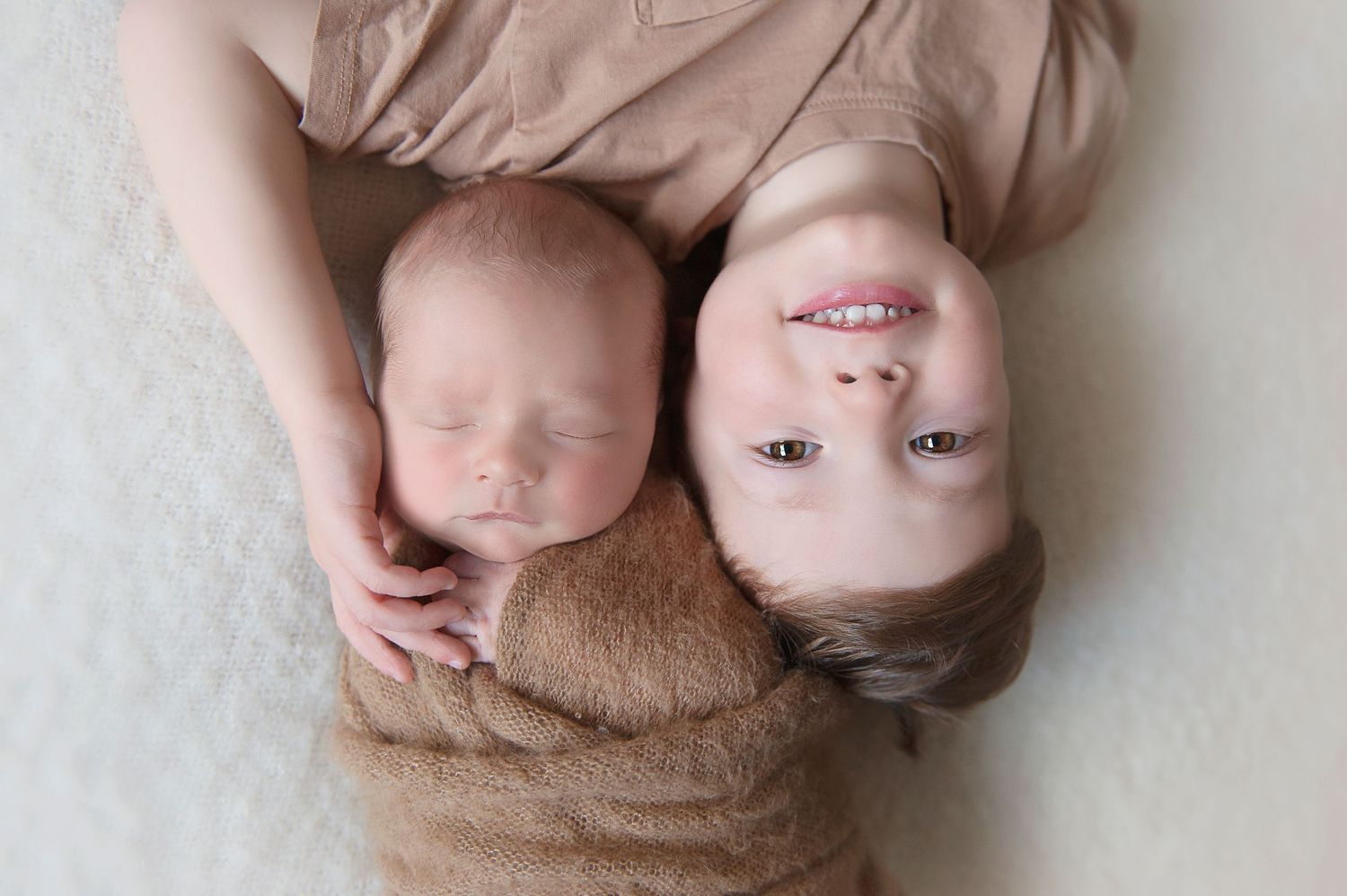 Overhead view of siblings lying on beige blanket during newborn photoshoot.