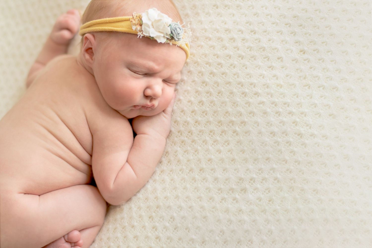 Sleeping newborn with delicate floral headband resting on textured cream surface.
