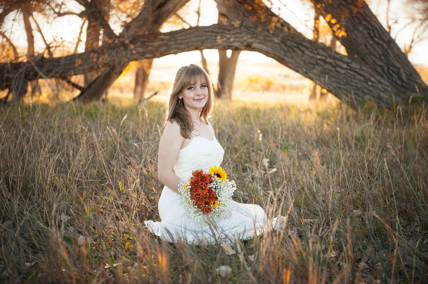Person in white dress sitting among tall grass with autumn trees behind.