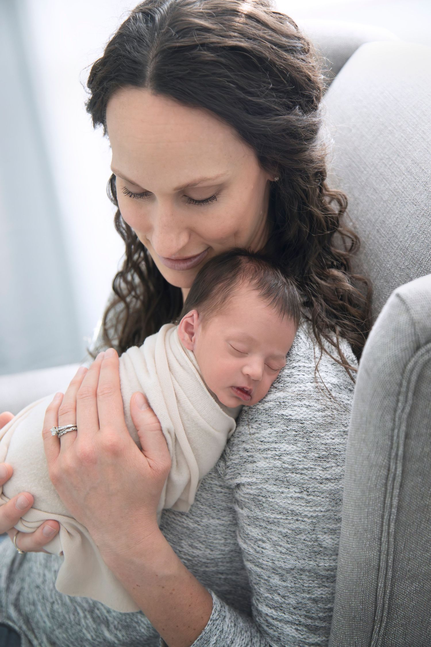 New parent cradling sleeping newborn baby while wearing grey sweater in soft natural light.