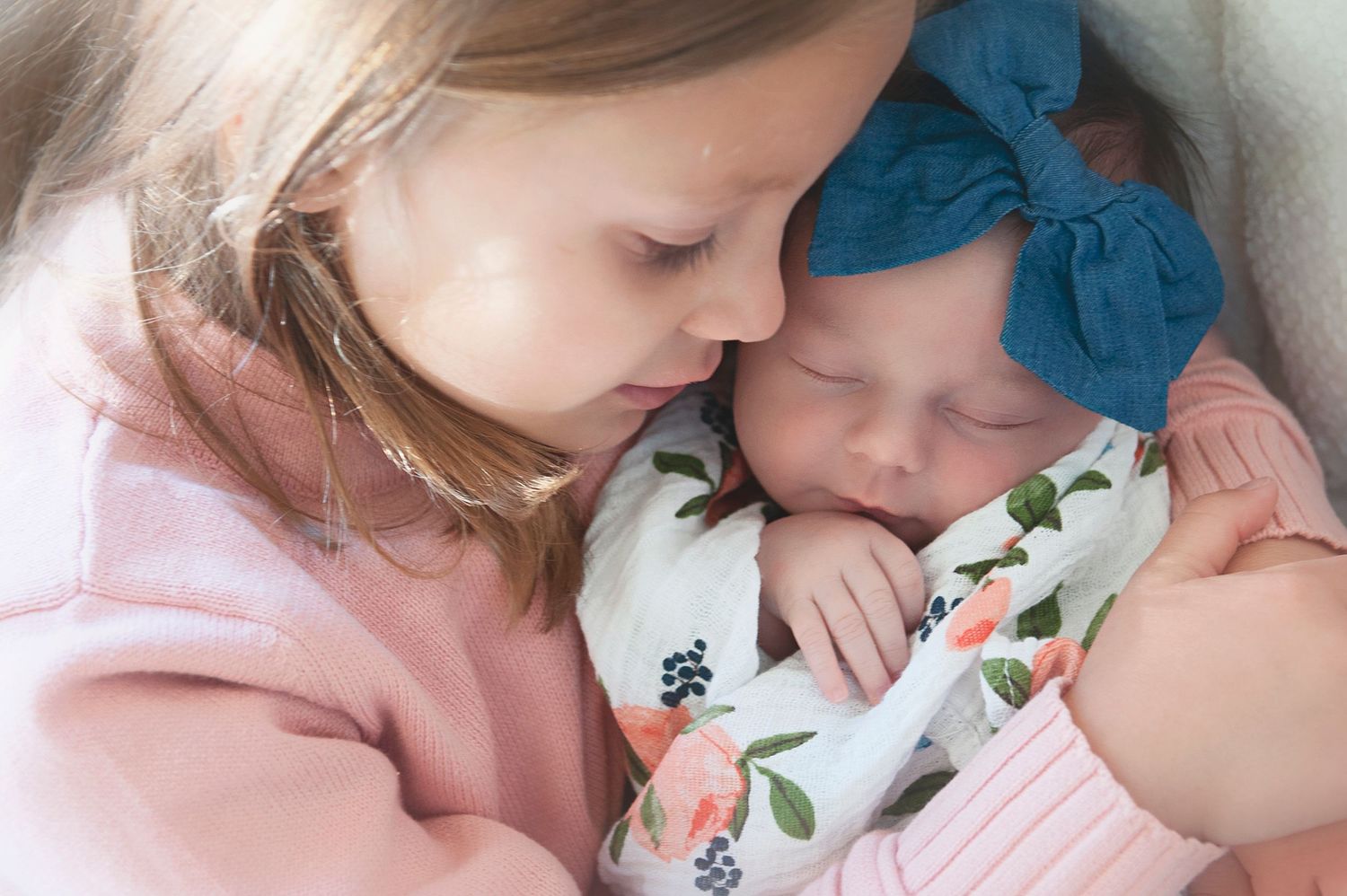 Two children share a tender moment in pink clothing with a floral swaddle and blue bow.