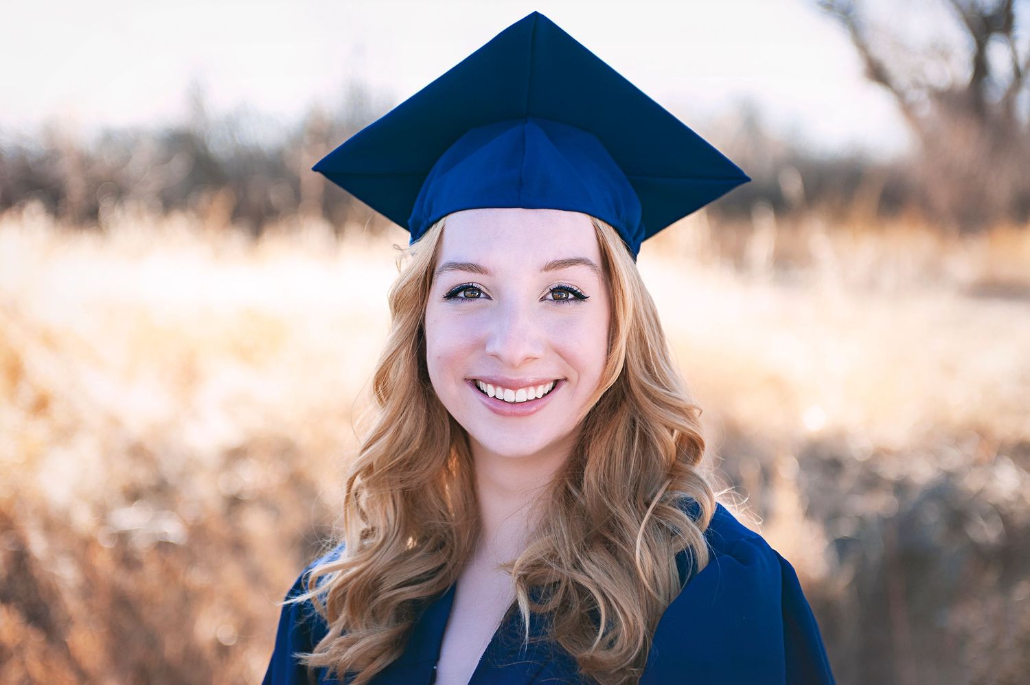 Graduate in navy blue cap and gown smiling against natural autumn background.