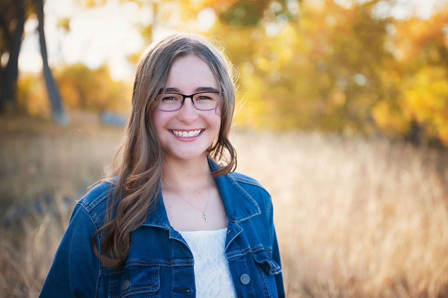 Person wearing glasses and denim jacket smiles warmly against golden autumn backdrop.