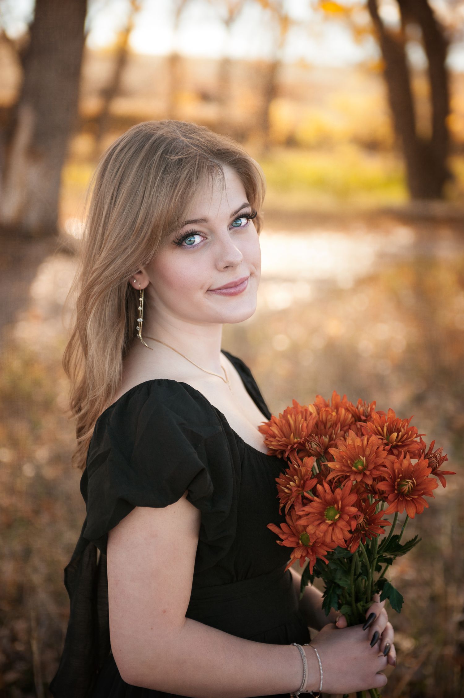 Portrait in black dress holding orange chrysanthemums with autumn trees in background.