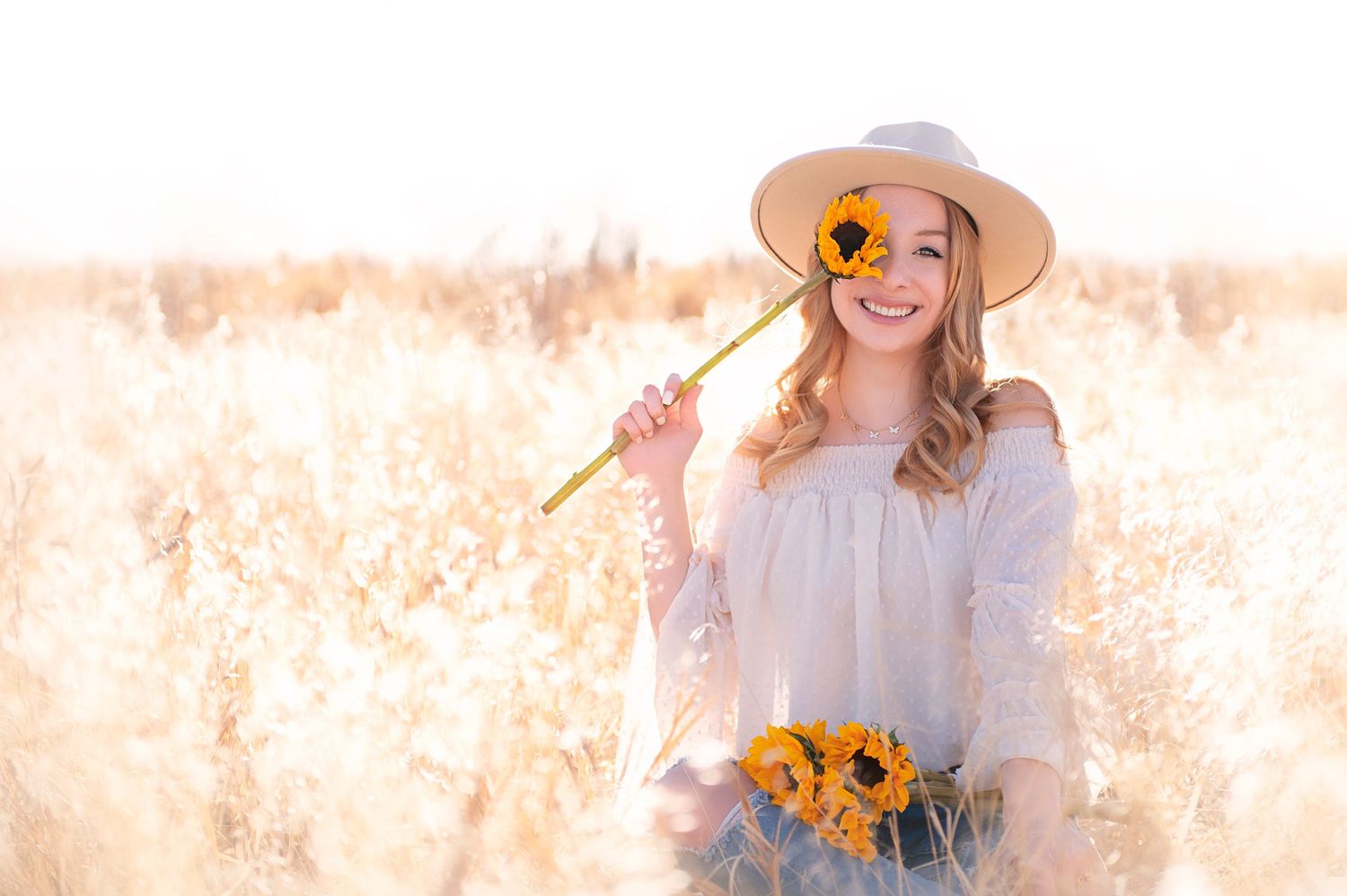 Playful pose with sunflower in wheat field wearing white dress and hat.