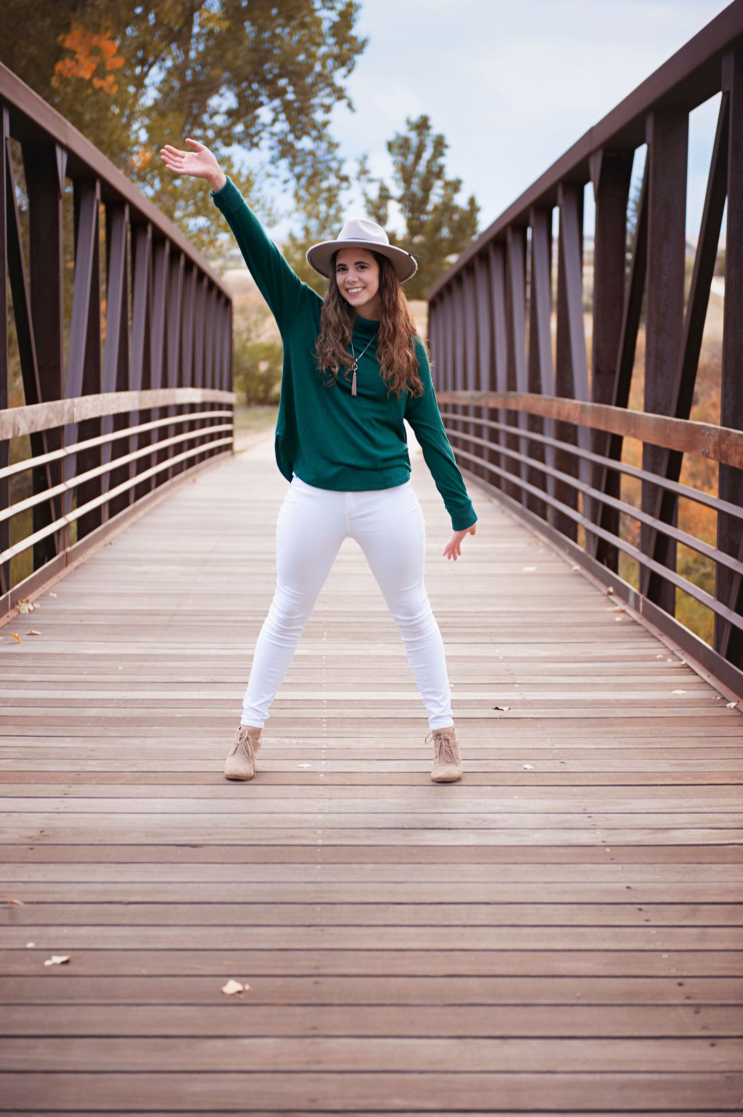 Person wearing green sweater and white pants strikes playful pose on wooden footbridge.
