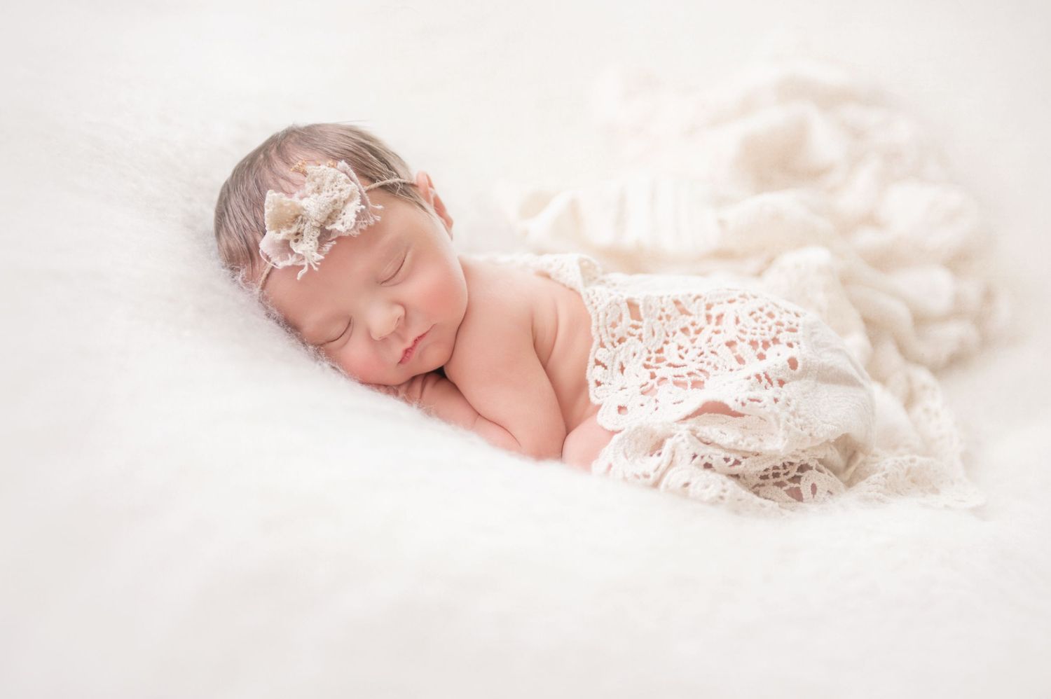Dreamy newborn portrait with lace wrap and floral headband on white backdrop.