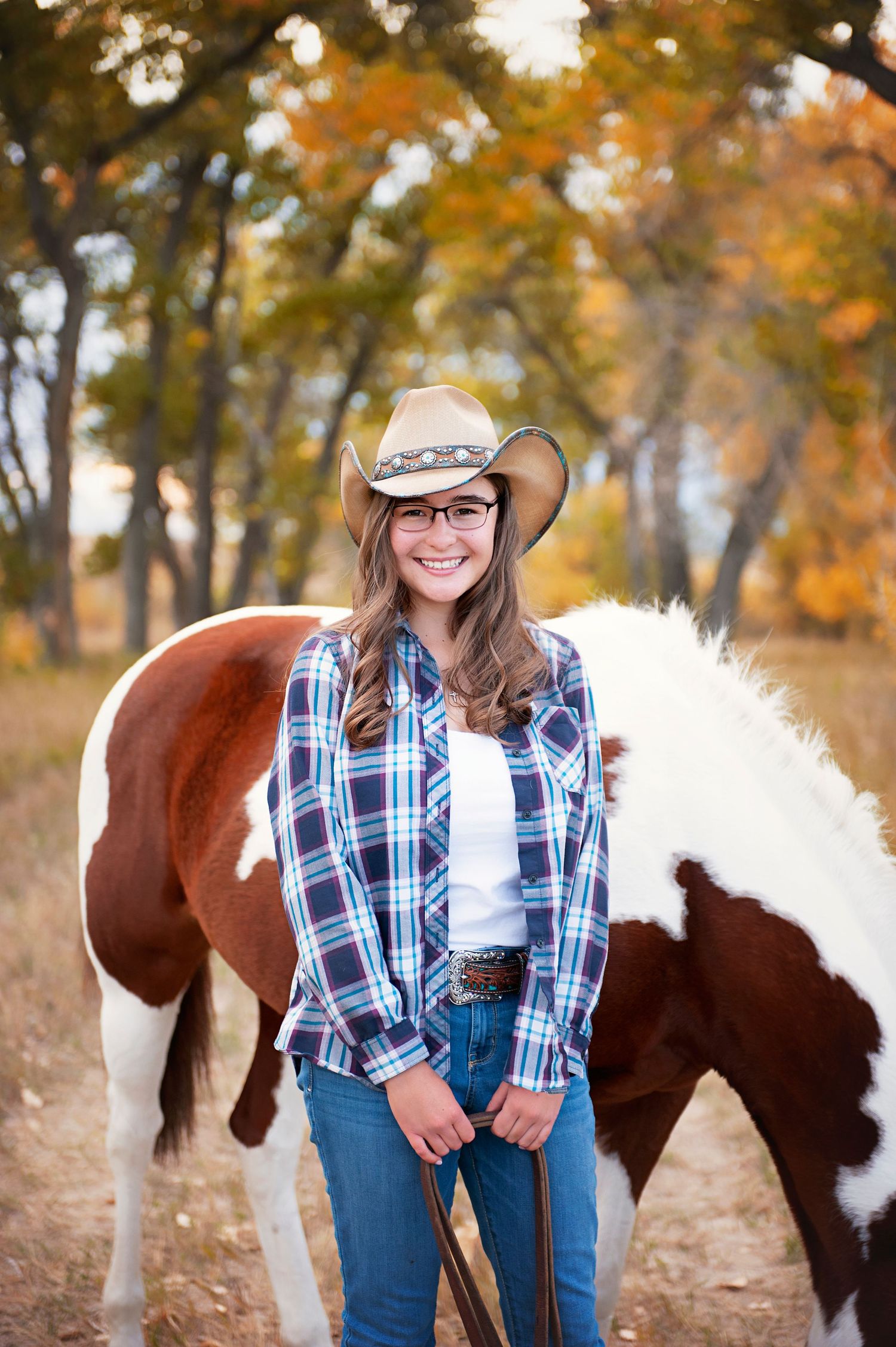 Person in western wear and cowboy hat poses with paint horse against fall foliage.