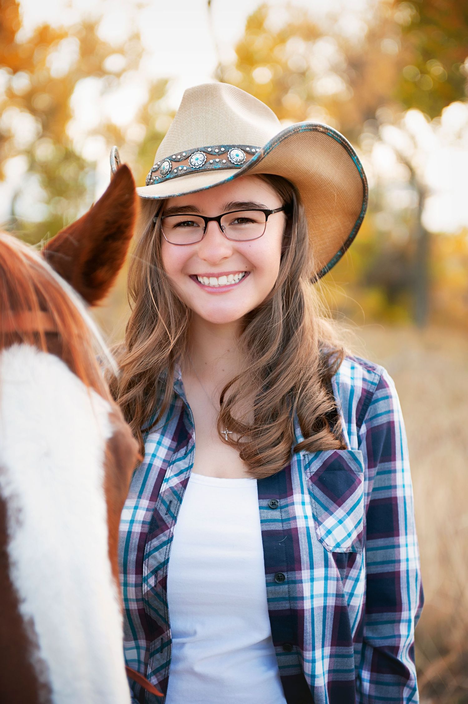 Western style portrait with plaid shirt and decorated cowboy hat against autumn foliage background.