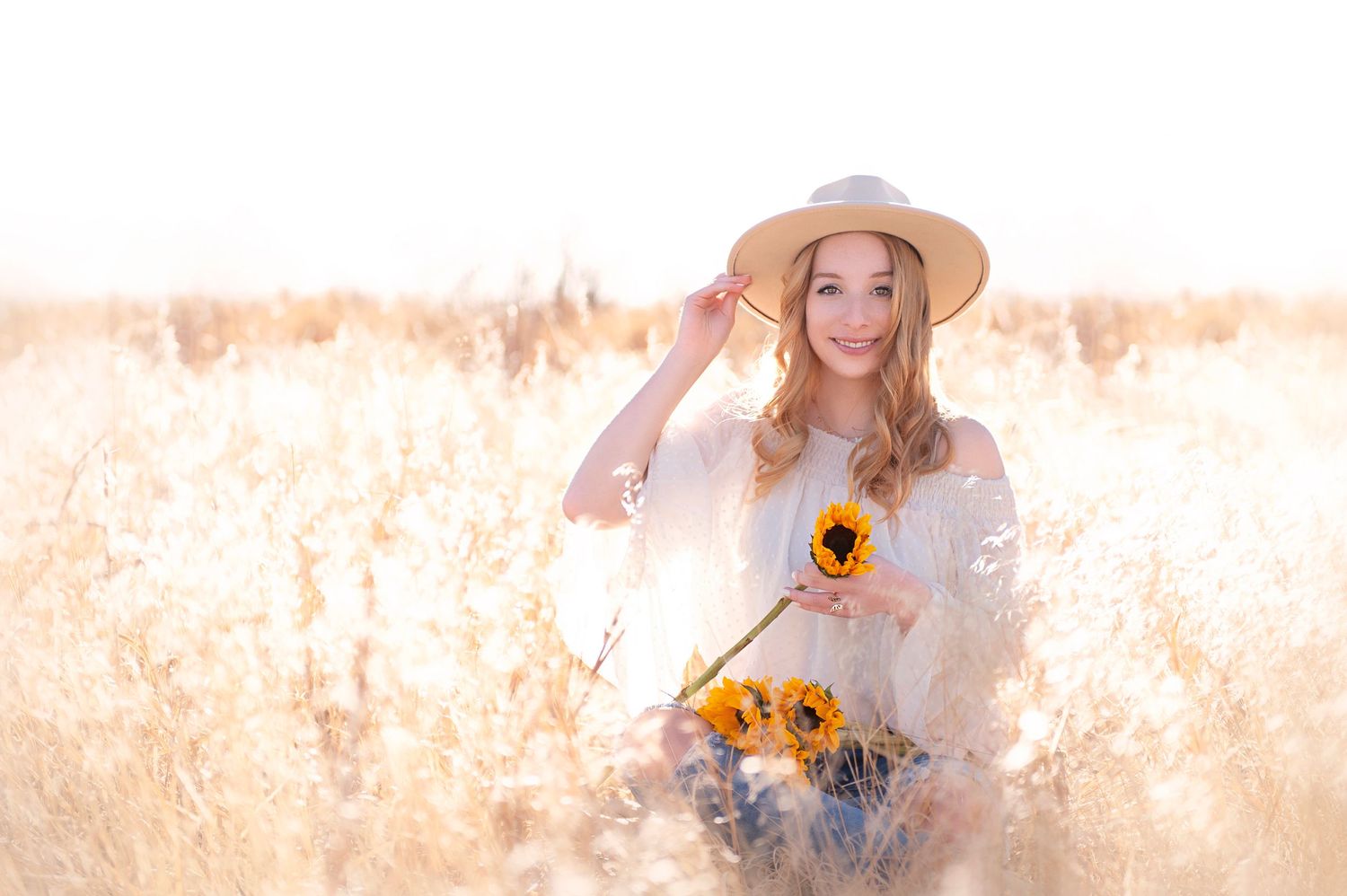 Person in a sun hat holding sunflowers in a golden wheat field during sunset.
