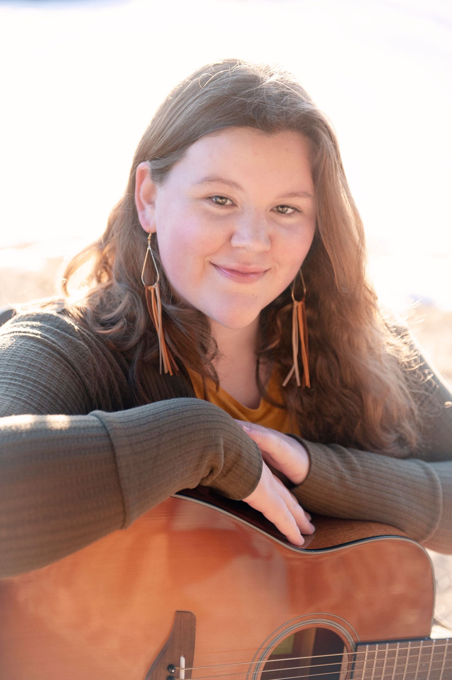 Person in gray sweater holding acoustic guitar with leather earrings against natural lighting.