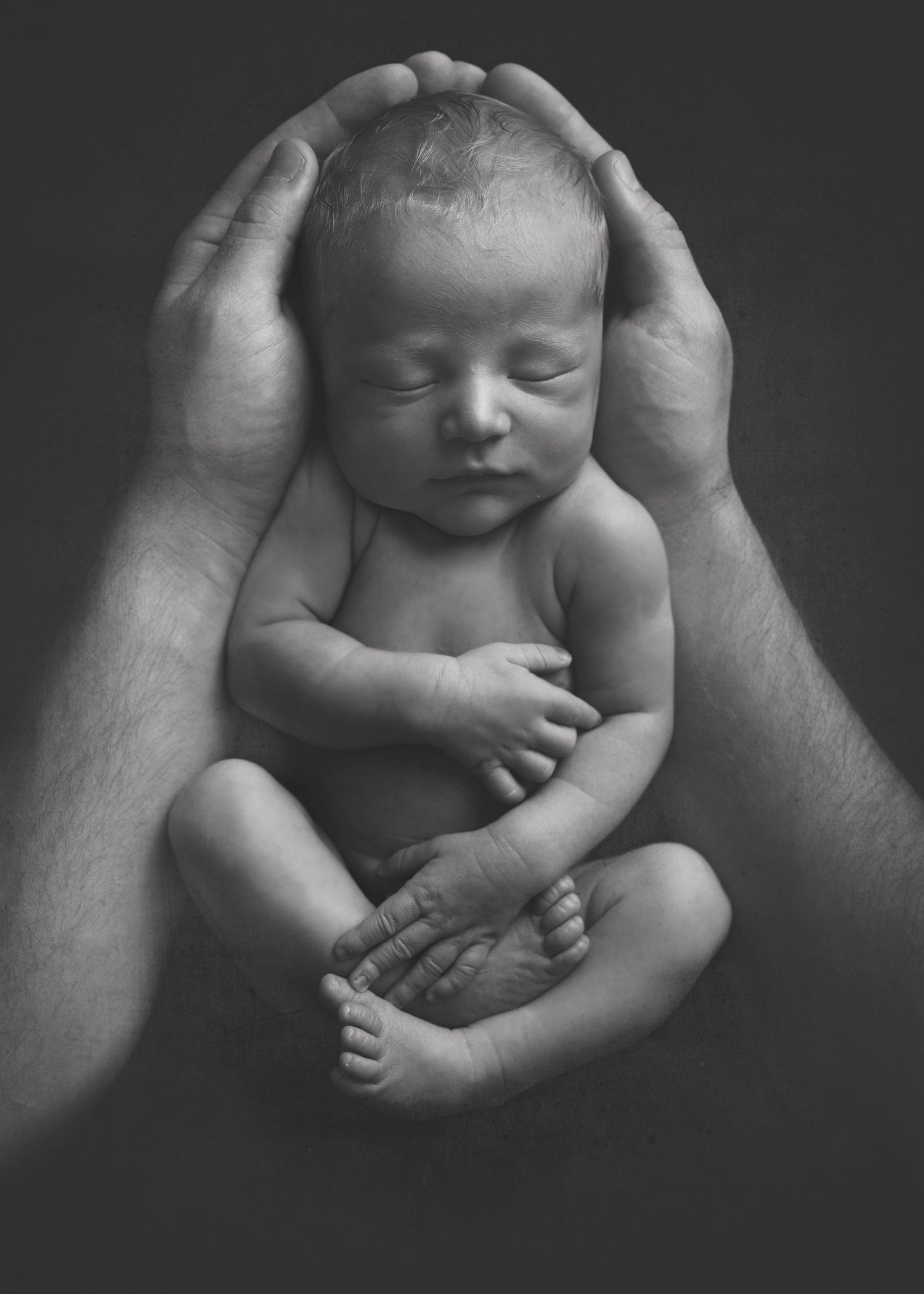 Black and white series of an infant sleeping peacefully in gentle supporting hands against a dark background.