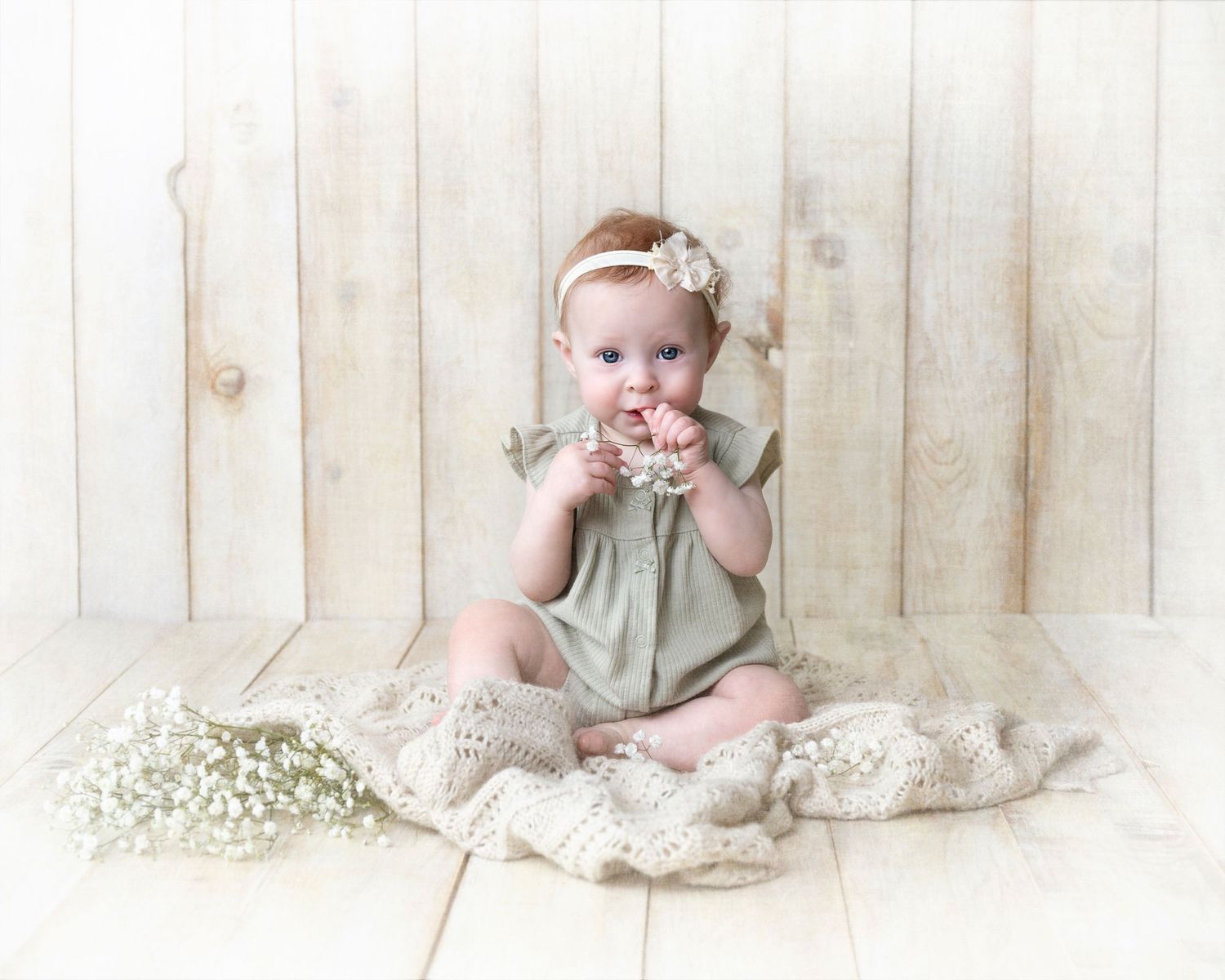 Small baby wearing sage green sleeveless dress sits on vintage lace with white flowers scattered around.