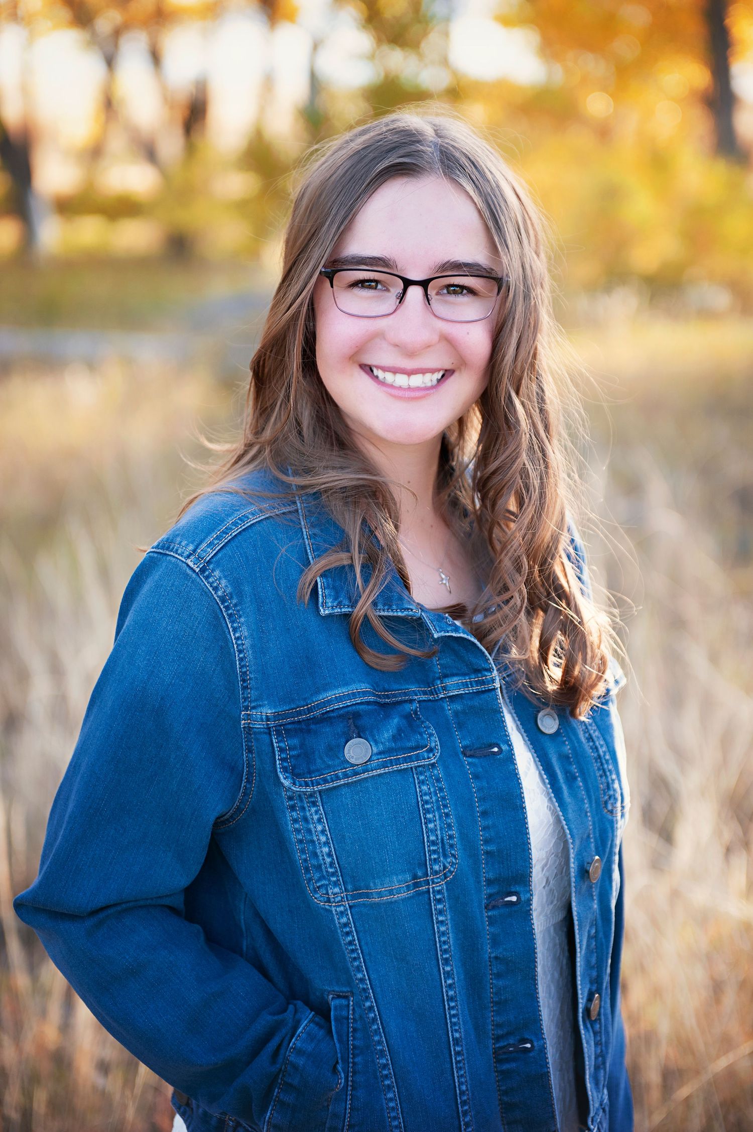 Portrait of someone in denim jacket and glasses with long hair against fall colors.
