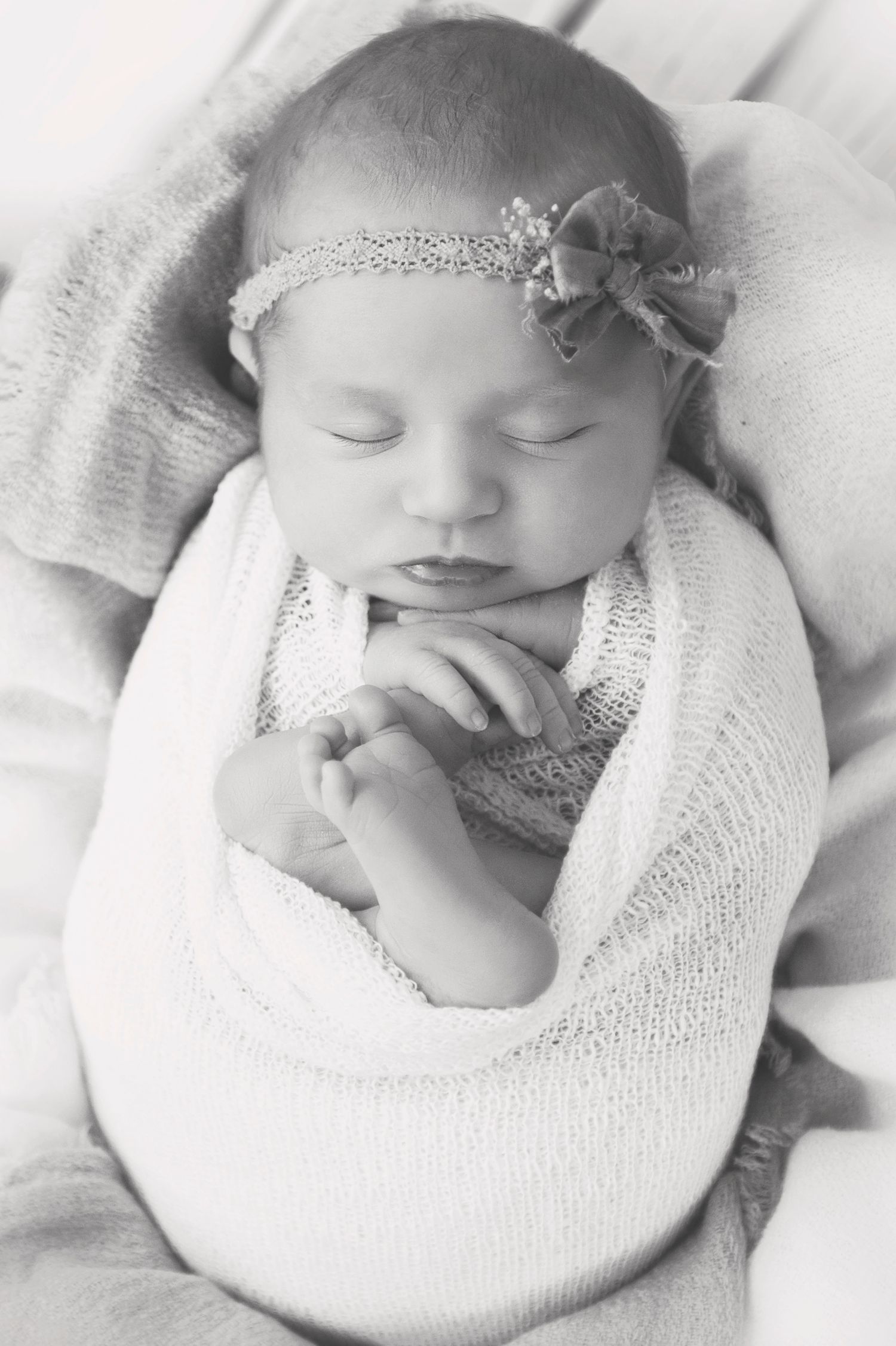 Peaceful newborn baby sleeps in white wrap wearing delicate headband in black and white portrait.
