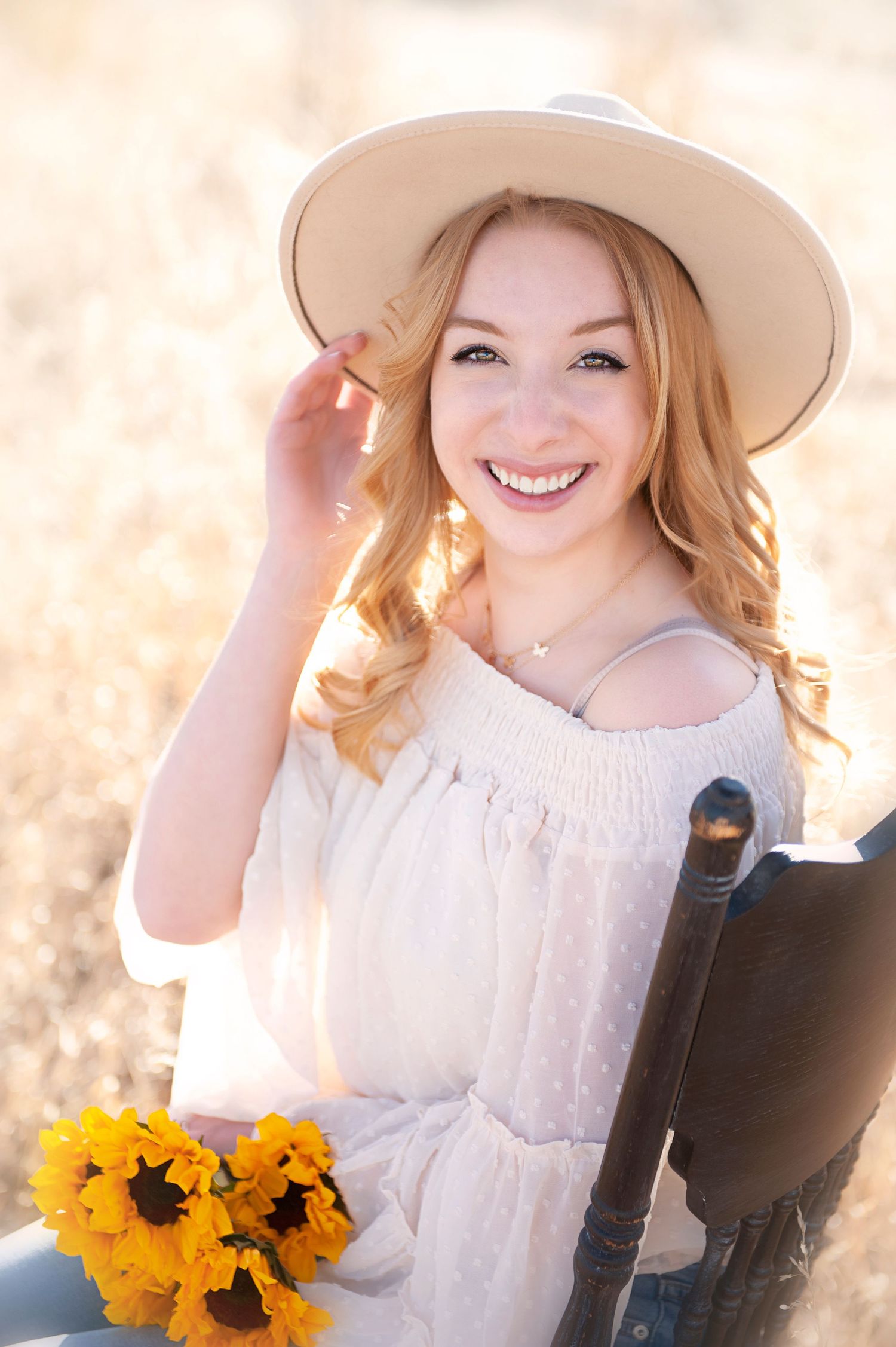 Person in white dress and beige hat poses with sunflowers in golden field.