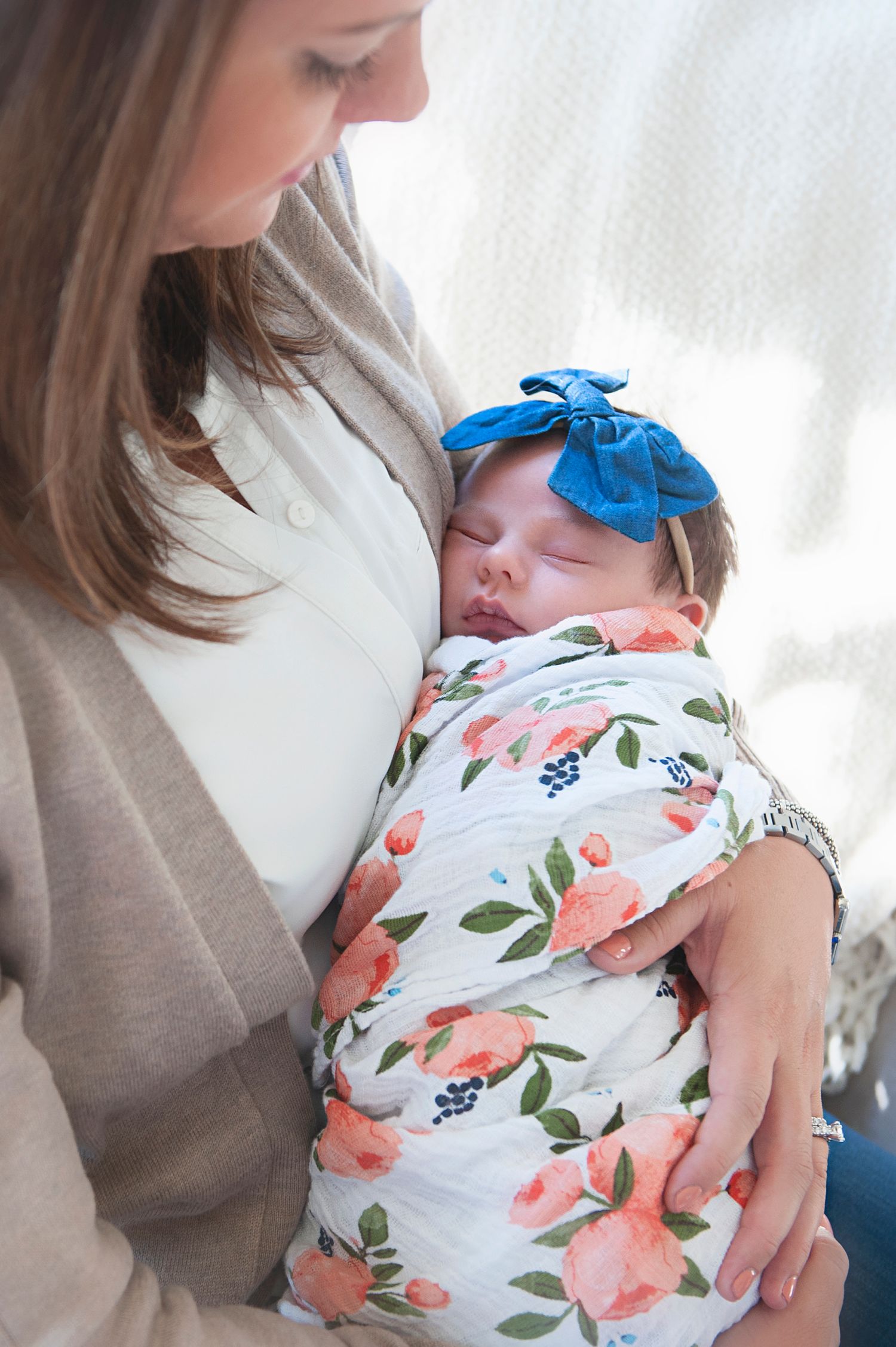 Infant in floral swaddle and blue bow rests against caregiver wearing neutral tones.
