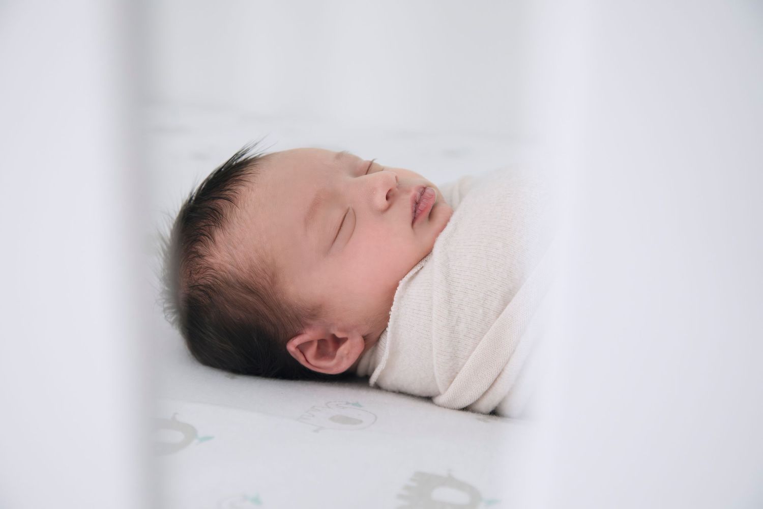 Peaceful sleeping newborn baby wrapped in cream colored swaddle against white bedding.
