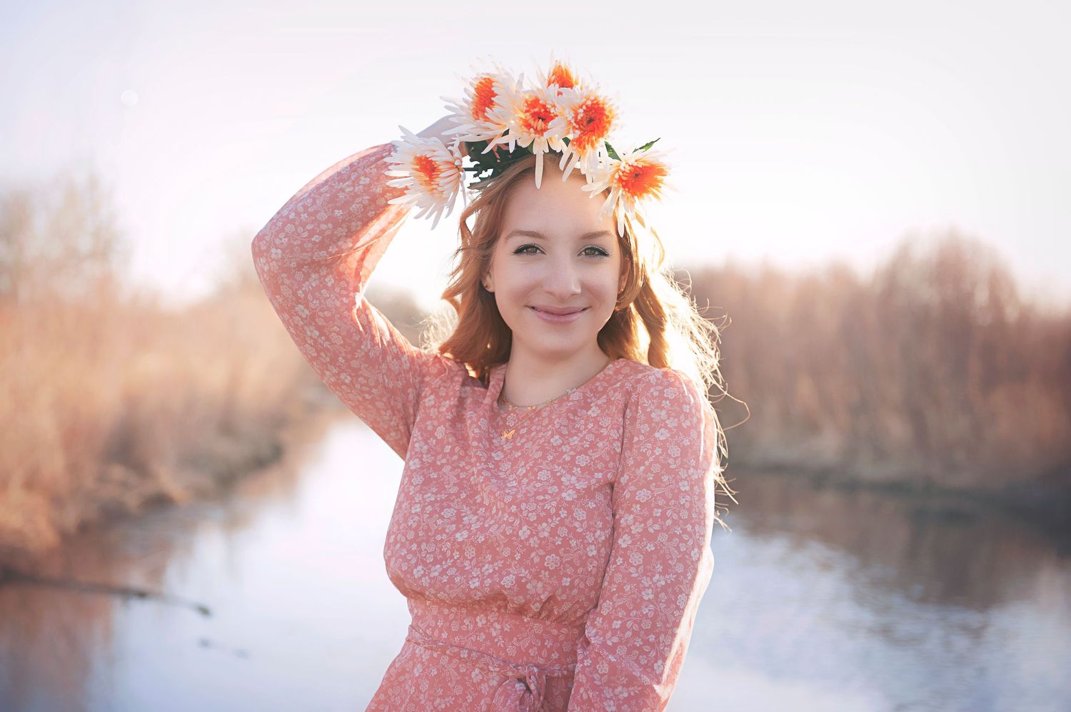 Person in pink dress wearing floral crown poses near water at golden hour.