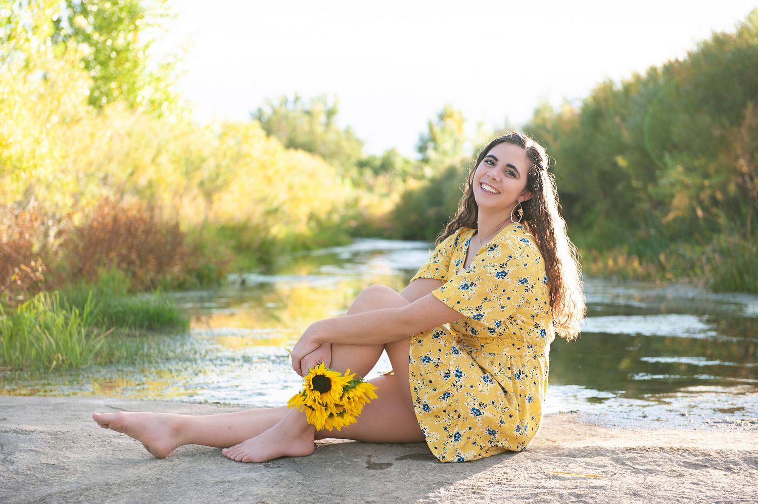 Person in yellow floral dress sitting barefoot by water's edge in summer setting.