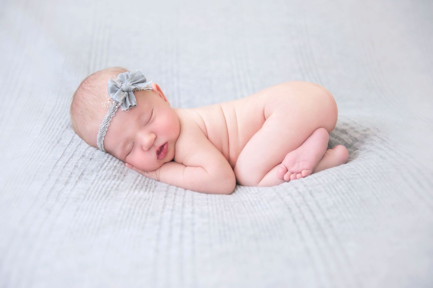 Sleeping newborn with gray bow headband lying on textured white blanket.