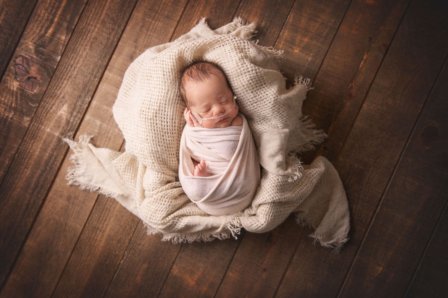 Newborn wrapped in white swaddle resting on cream textured blanket over wooden floor.