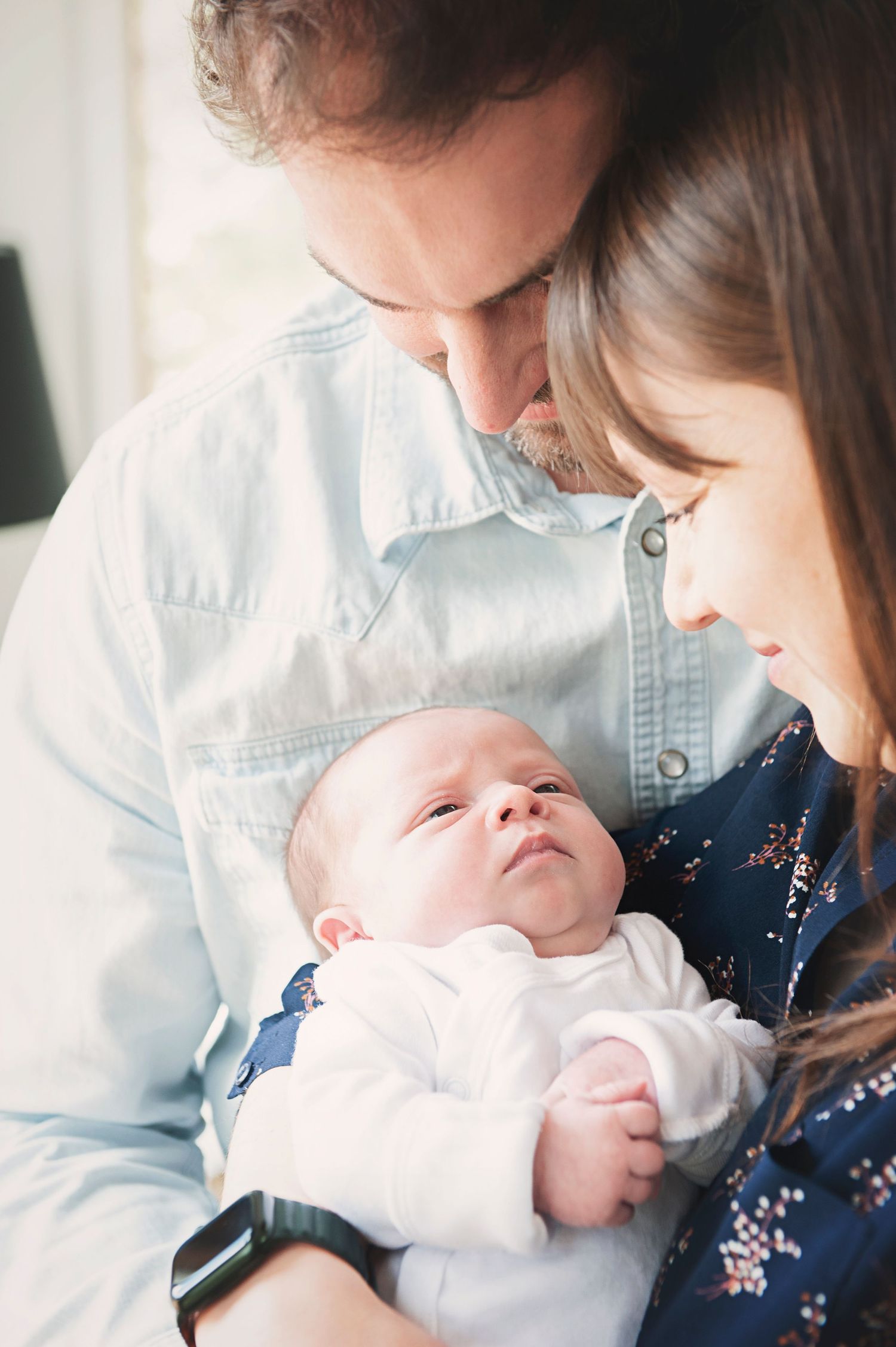 Parents in casual attire gazing lovingly at their alert newborn baby between them.
