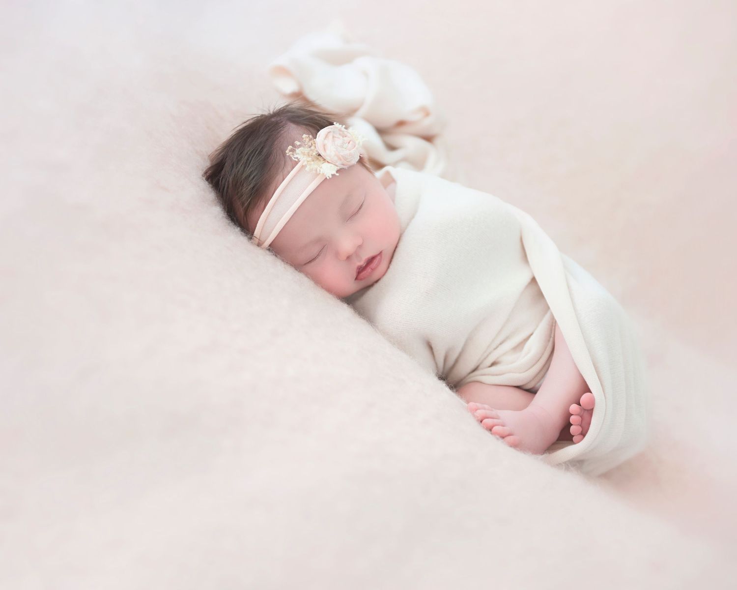 Newborn baby sleeping peacefully in white wrap with delicate headband on pink background.
