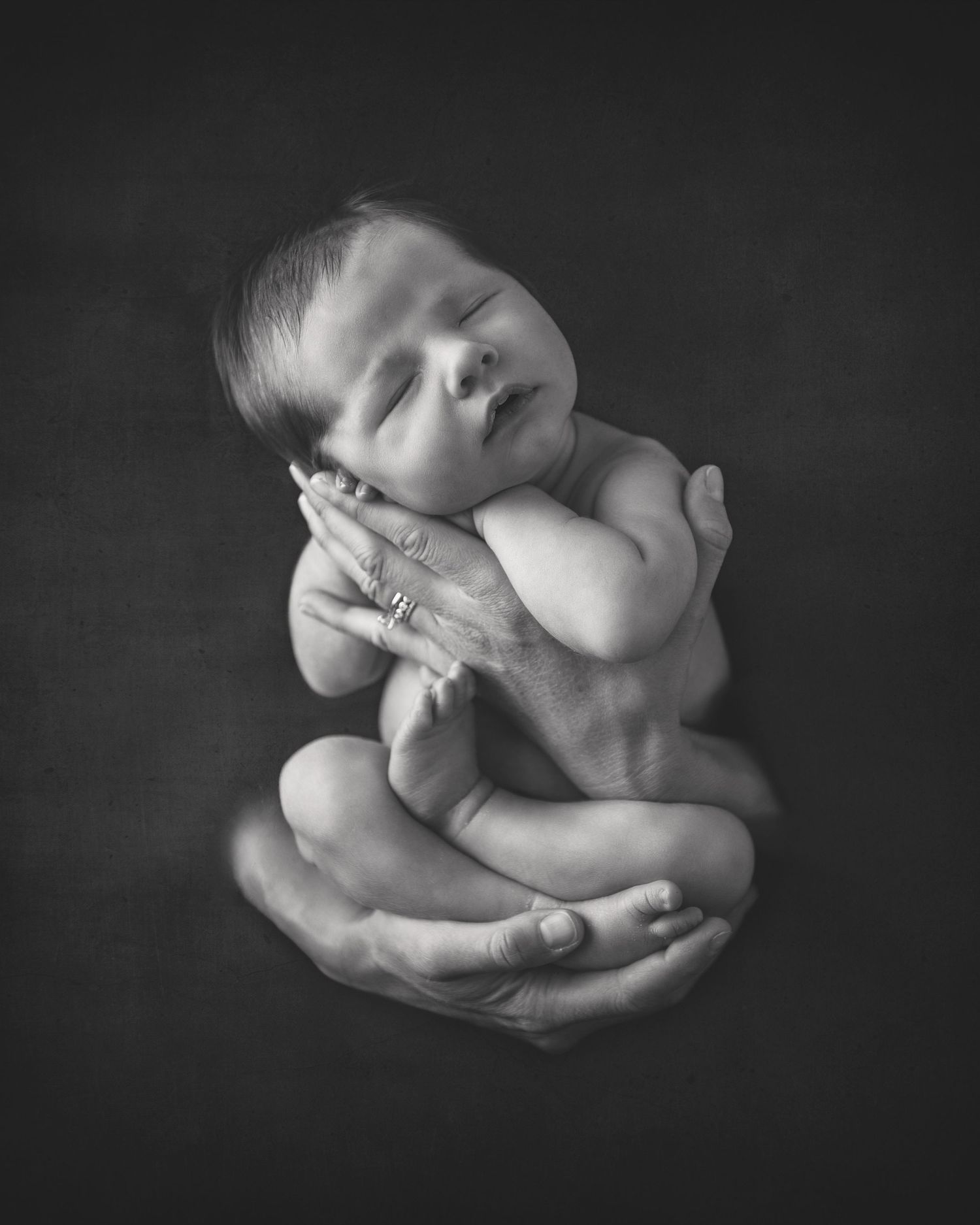Artistic black and white portrait of newborn cradled in adult hands against dark background.