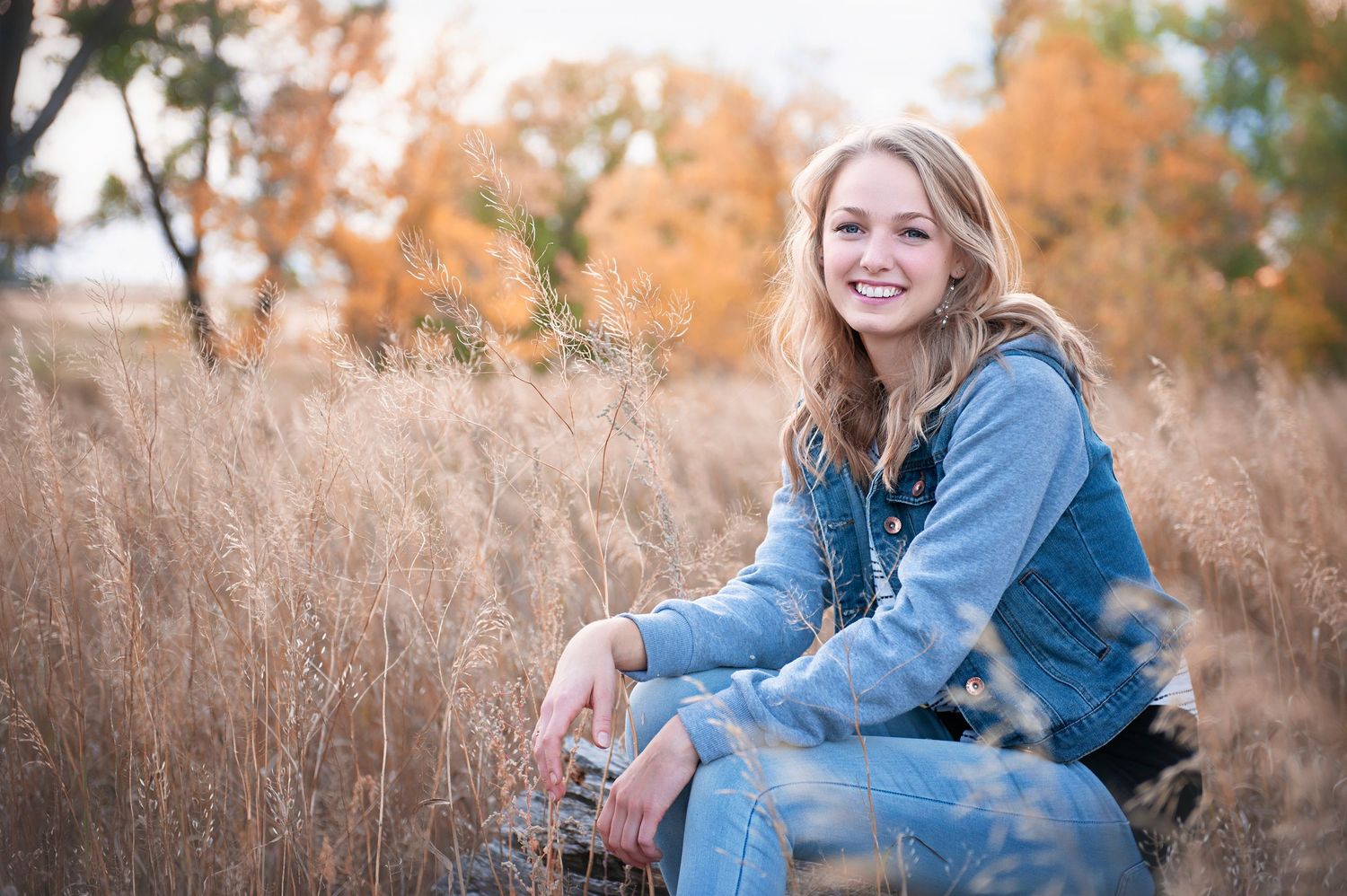 Casual pose in denim outfit sitting among tall autumn grass.