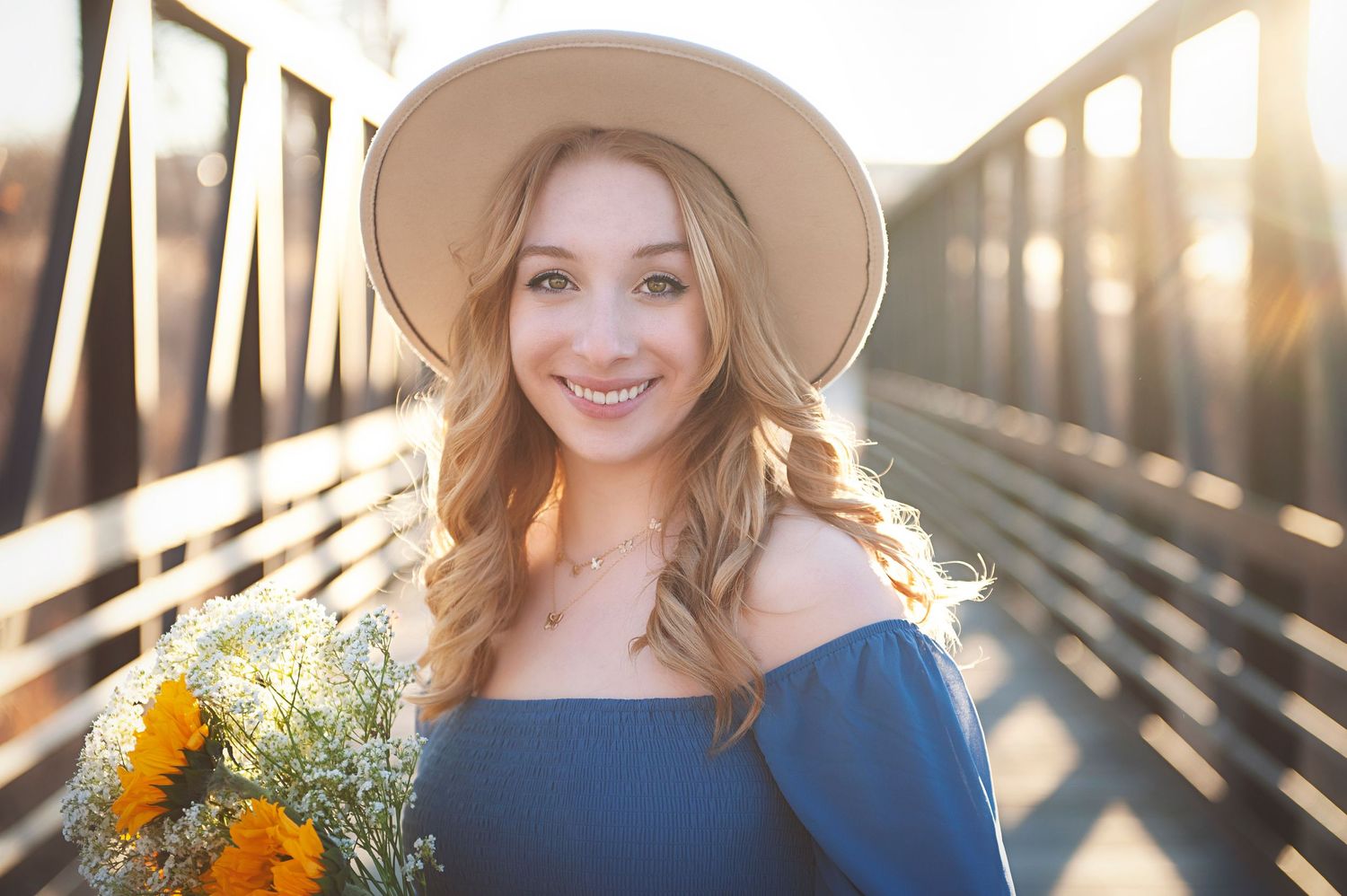 Golden hour portrait on bridge with blue dress, hat and sunflower bouquet.