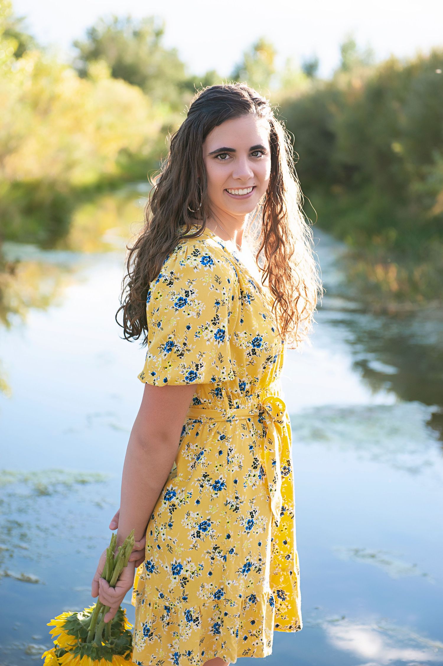Person in yellow floral dress holding flowers by waterside in summer setting.