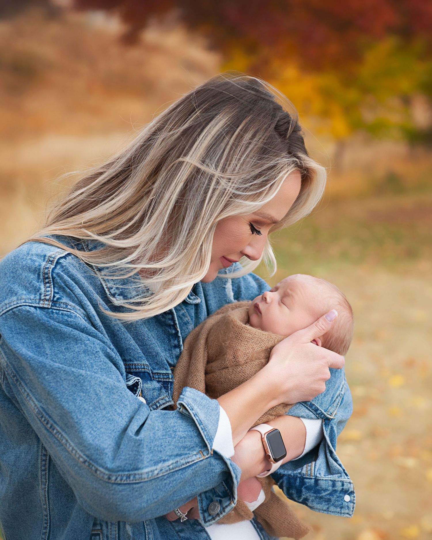 A woman in denim cradles a baby wrapped in brown against an autumn background.