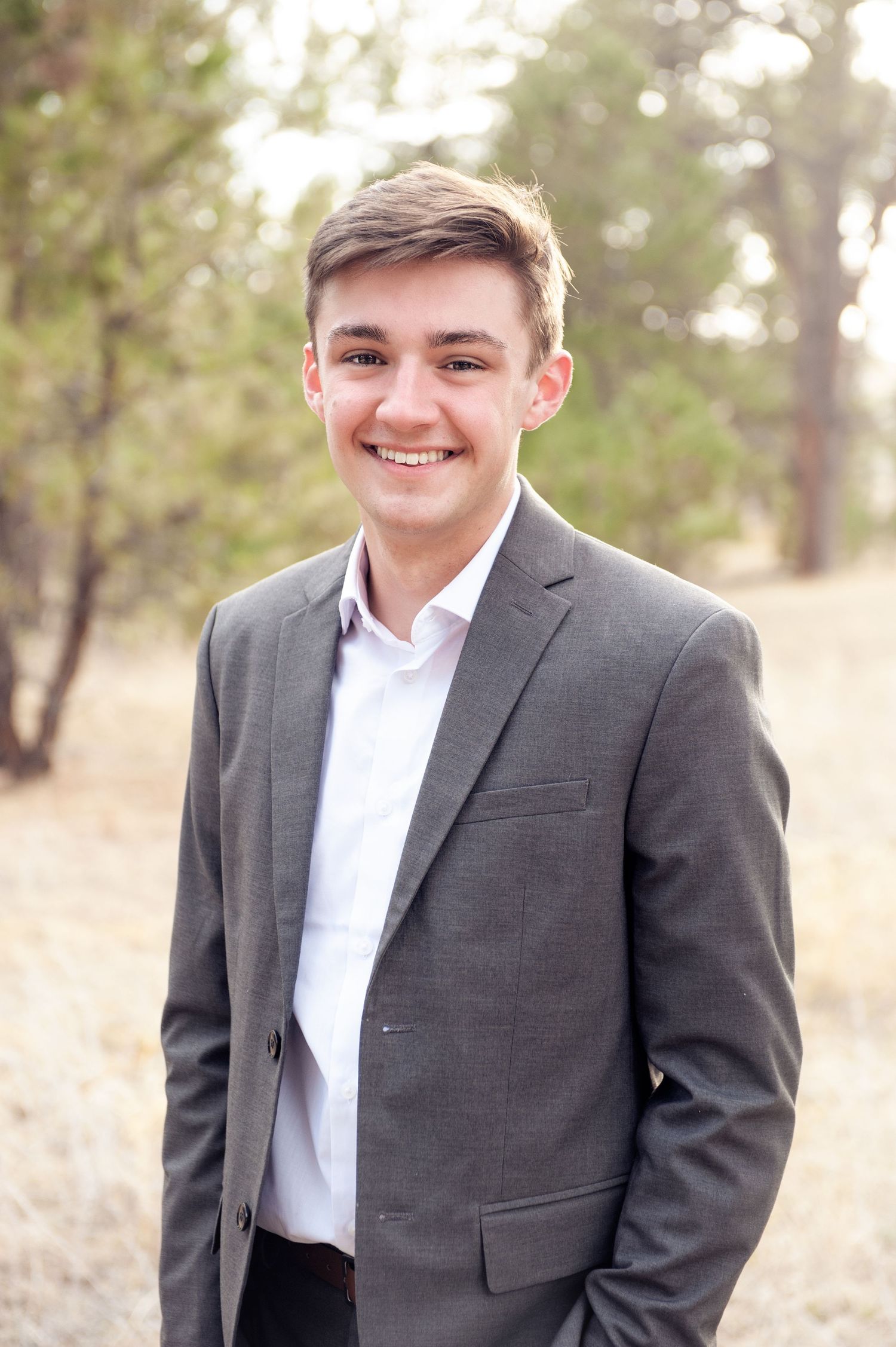Person in gray suit and white dress shirt standing in front of pine trees with natural lighting.