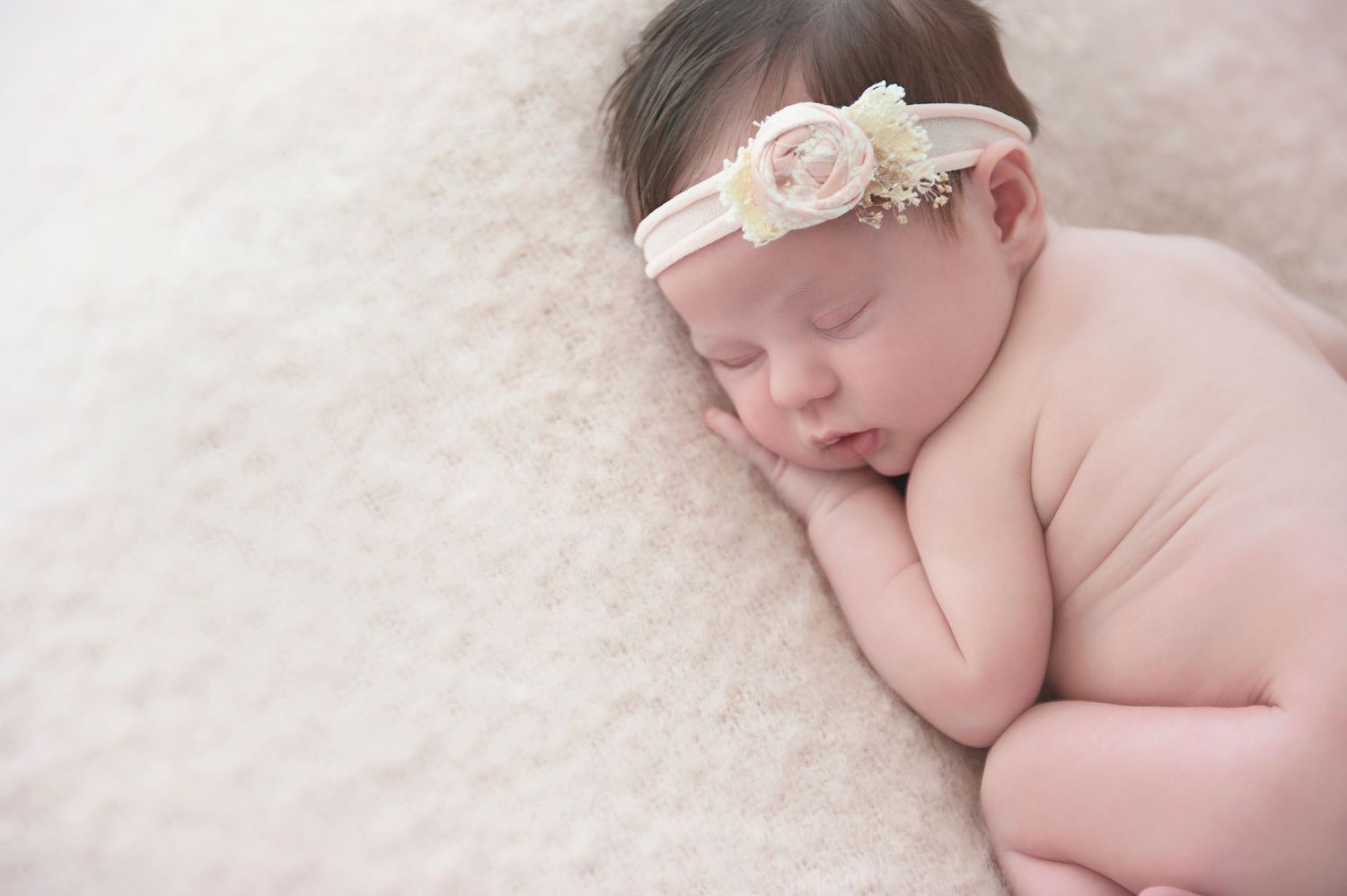 Serene sleeping newborn wearing pink floral headband resting on plush cream blanket.