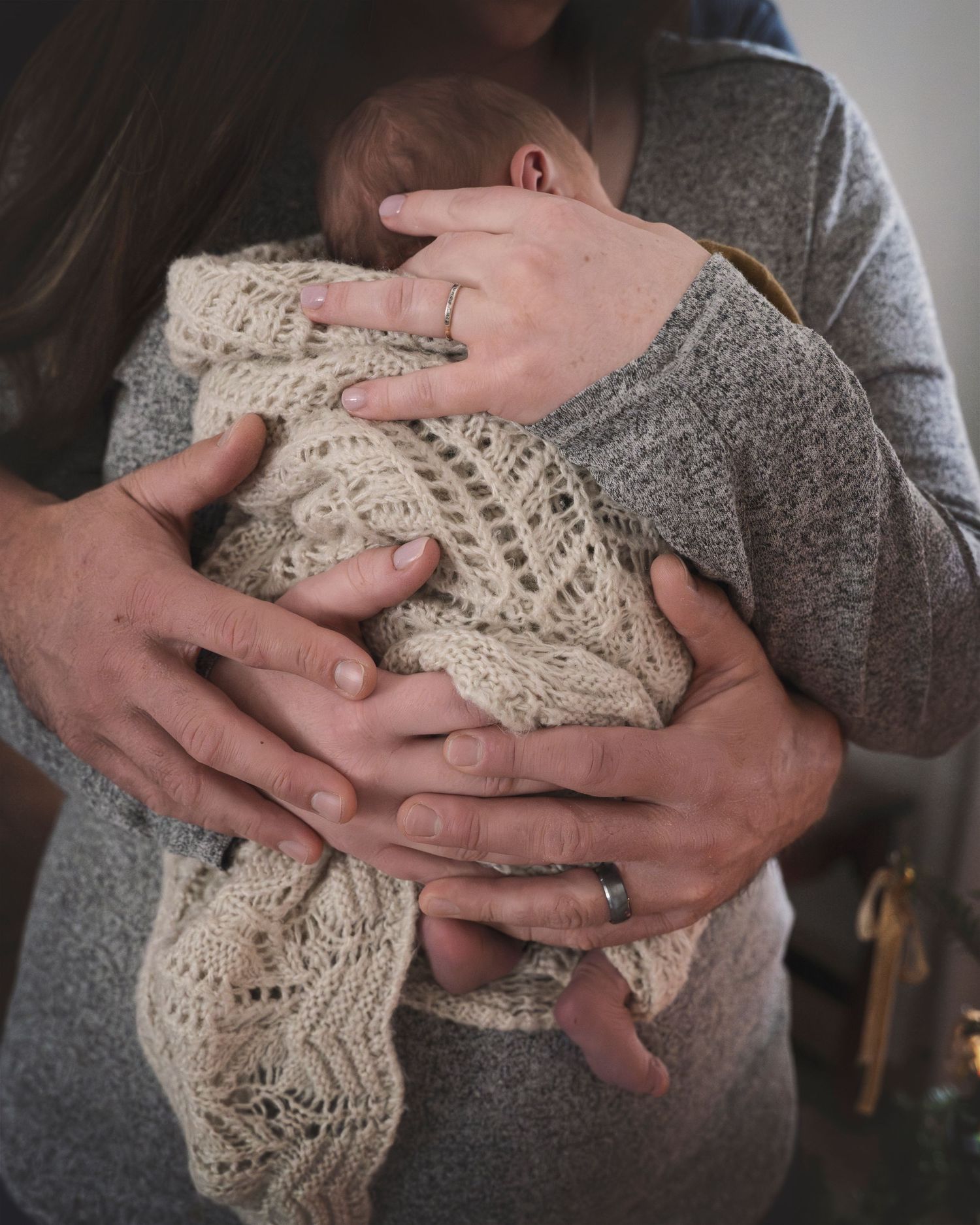 A tender moment of hands cradling a newborn wrapped in a knit cream-colored blanket.