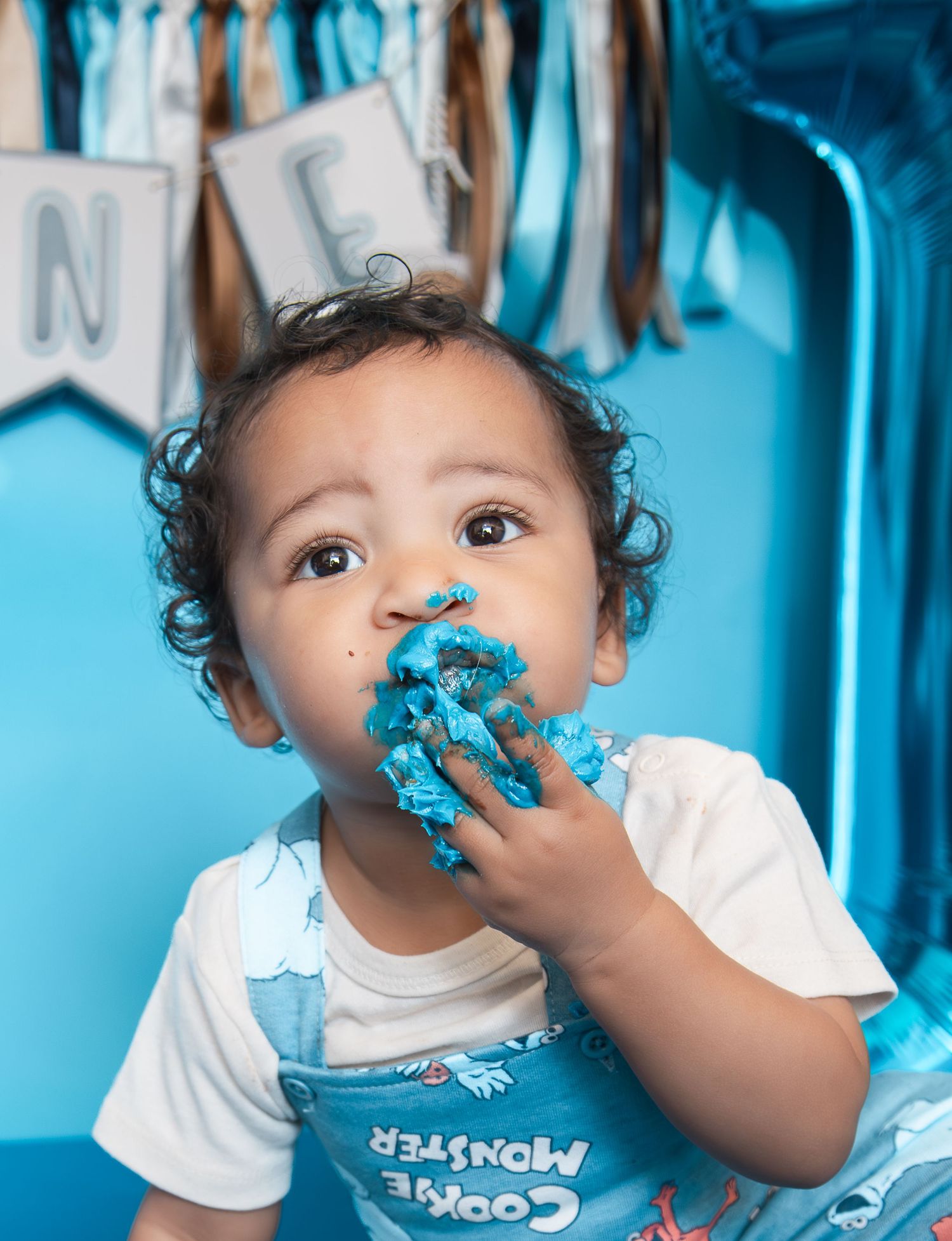 Baby in Cookie Monster bib enjoys first birthday cake smash with blue frosting on face and hands.