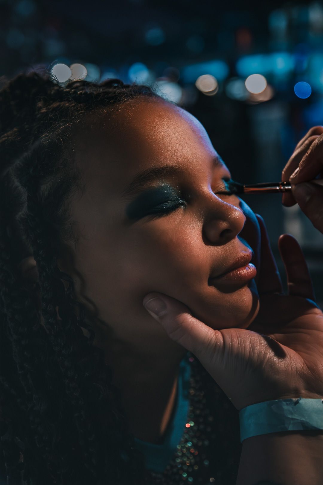 A makeup artist applies teal eyeshadow to a young Black woman with curly hair under moody blue studio lighting.