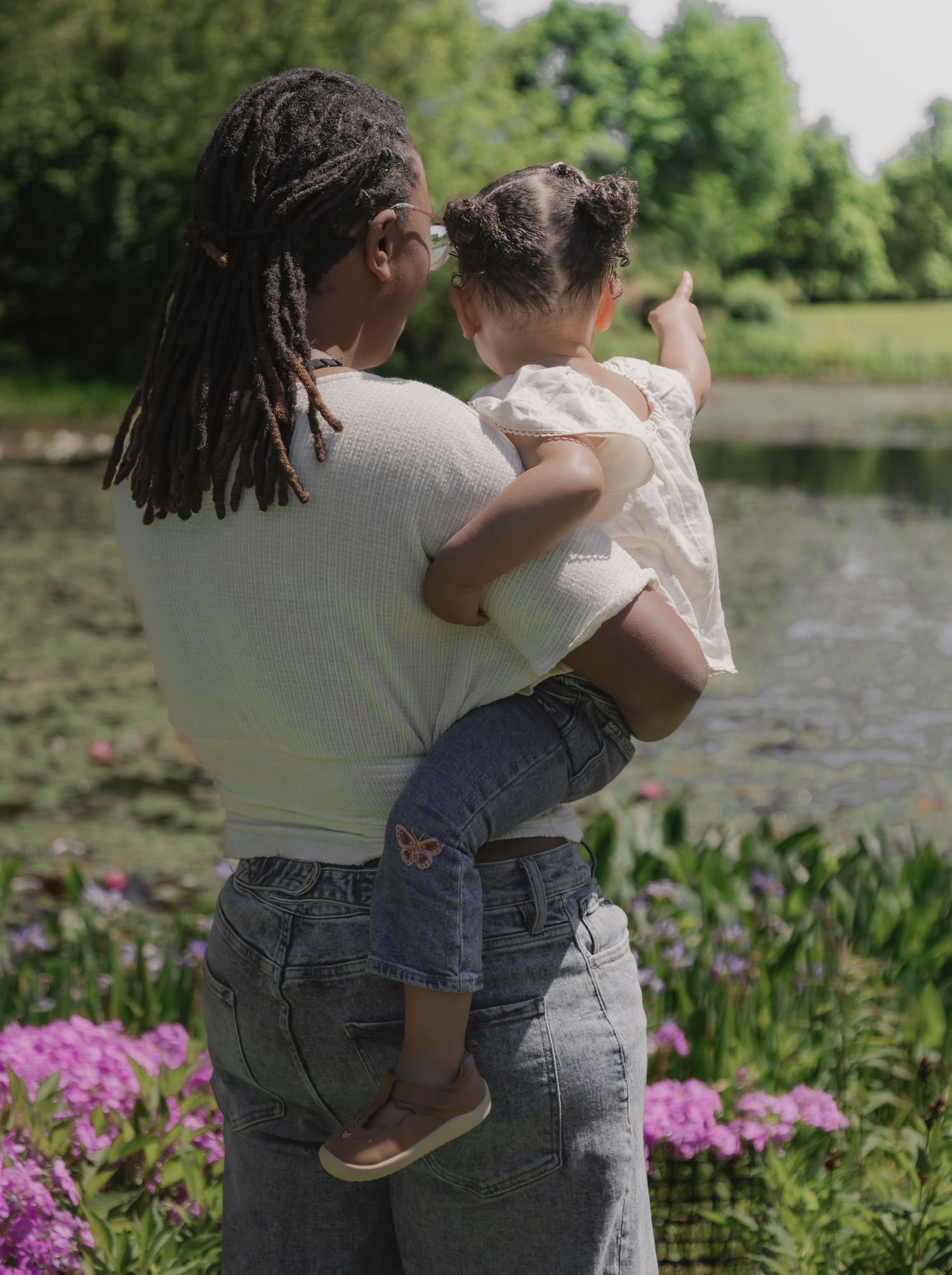 A mother holds her toddler while standing by a serene pond surrounded by lush greenery and vibrant pink flowers.
