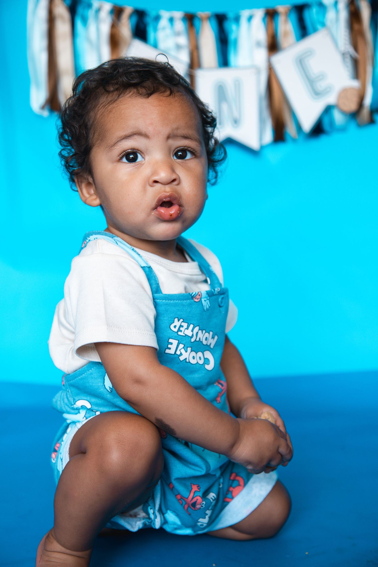 Cute baby boy in Cookie Monster overalls sitting on blue floor at his first birthday photo shoot.