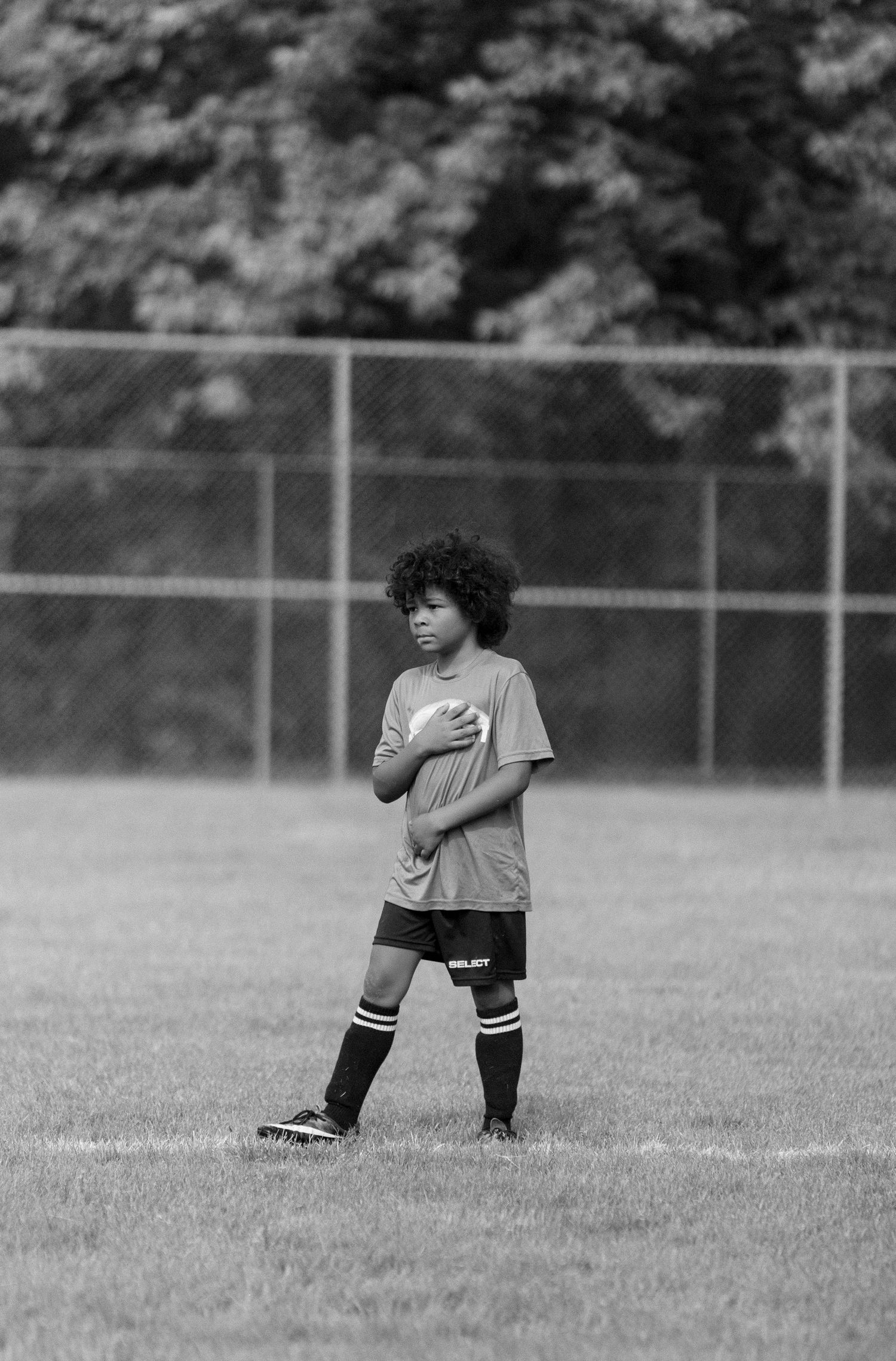 Black and white photo of a young curly-haired boy in soccer uniform standing on a grass field.
