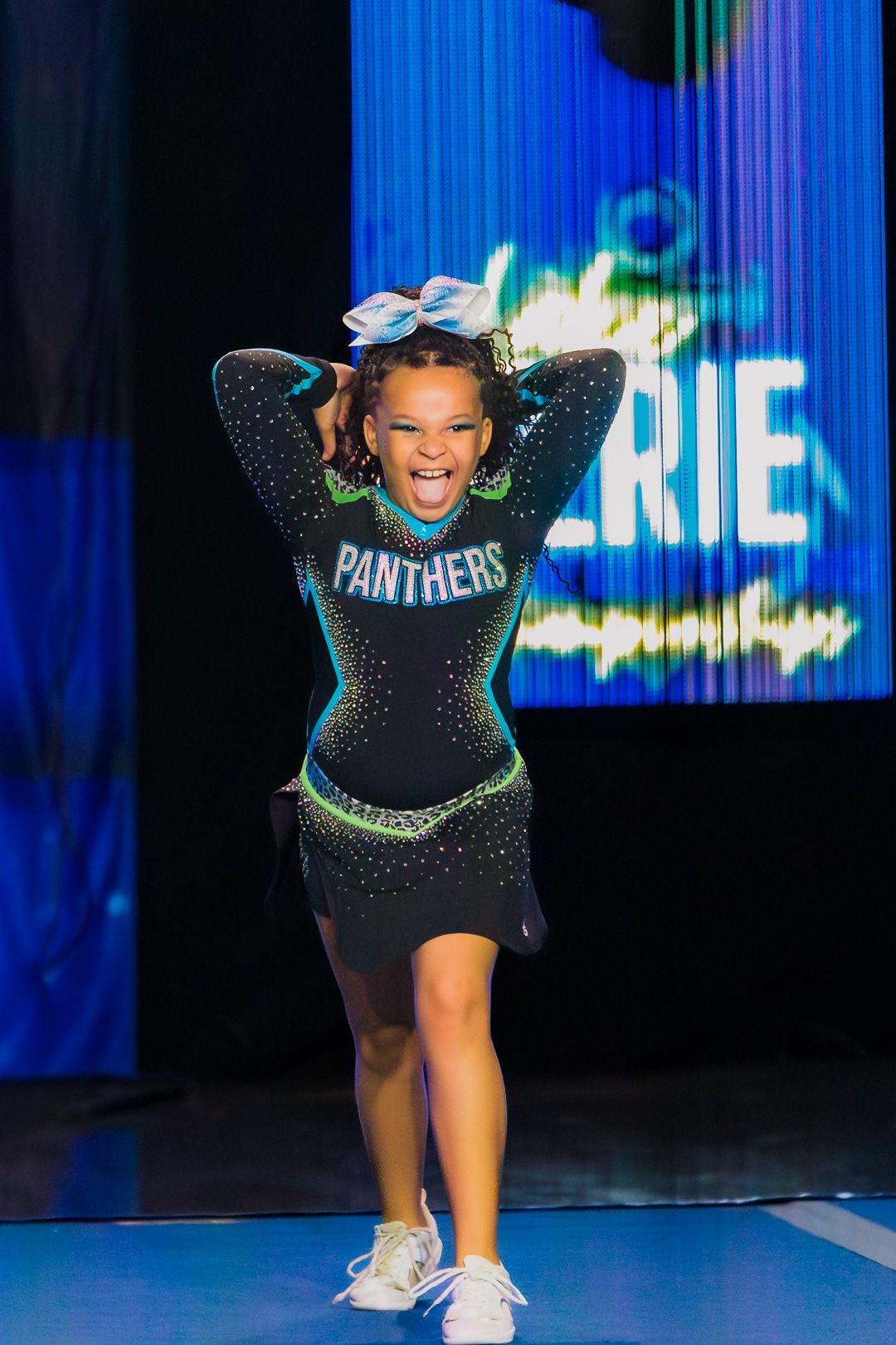 Young cheerleader in Panthers uniform performs energetically on stage at a cheer competition.