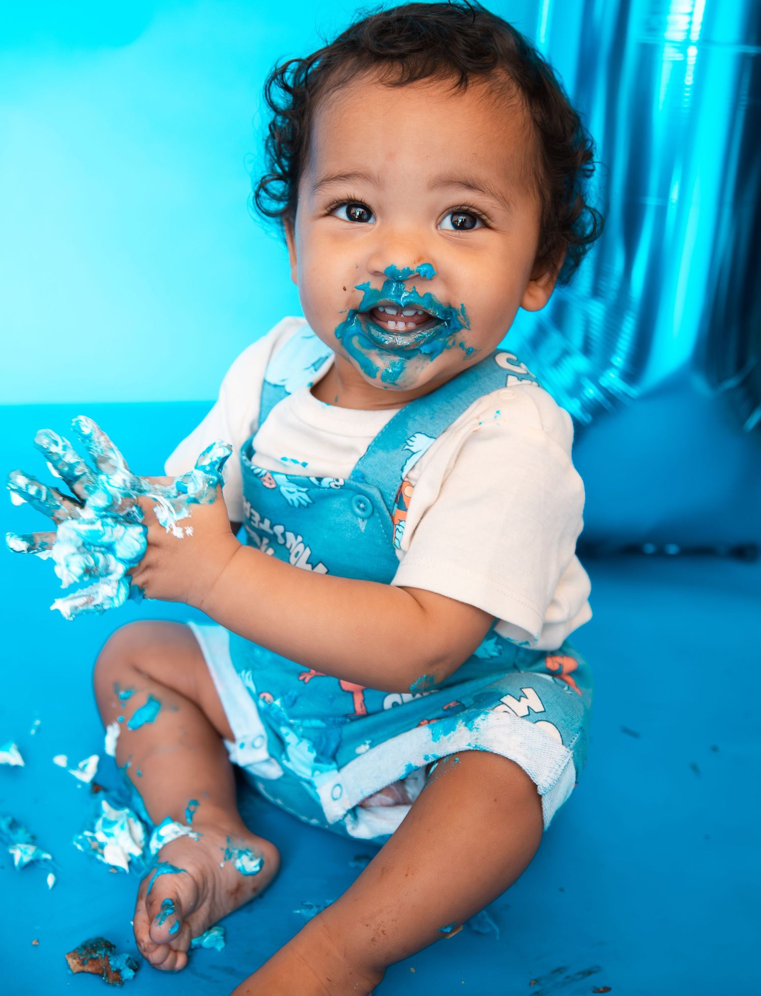 Baby in blue overalls smiling with blue cake smash frosting on face and hands during first birthday photo shoot.
